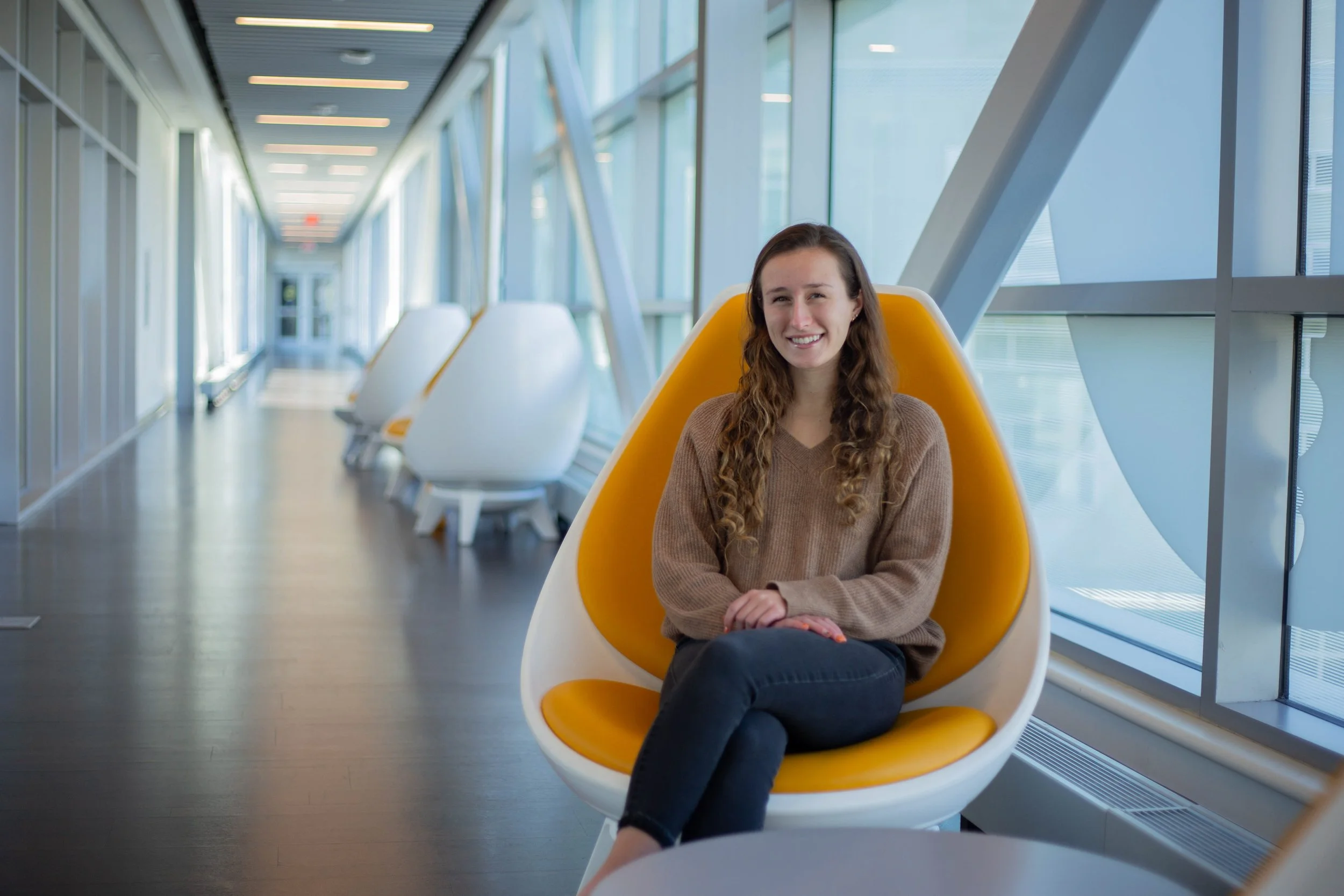 A young woman with long, curly brown hair sitting in a modern airport lounge chair, smiling at the camera. The lounge has large windows letting in natural light, and a row of empty white and yellow chairs is visible along the corridor.