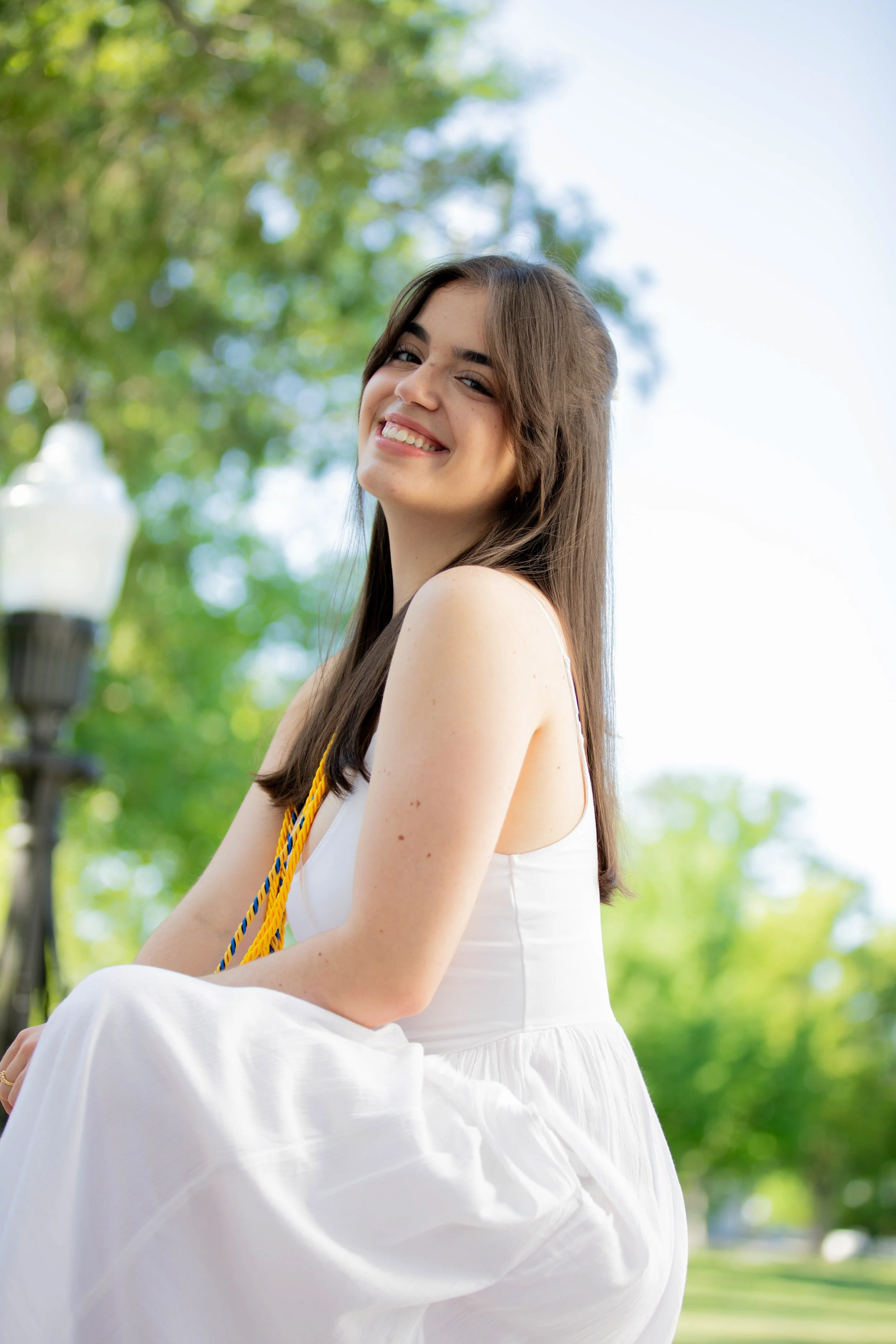 Young woman smiling outdoors in a white dress, sitting with green trees and blue sky in the background.