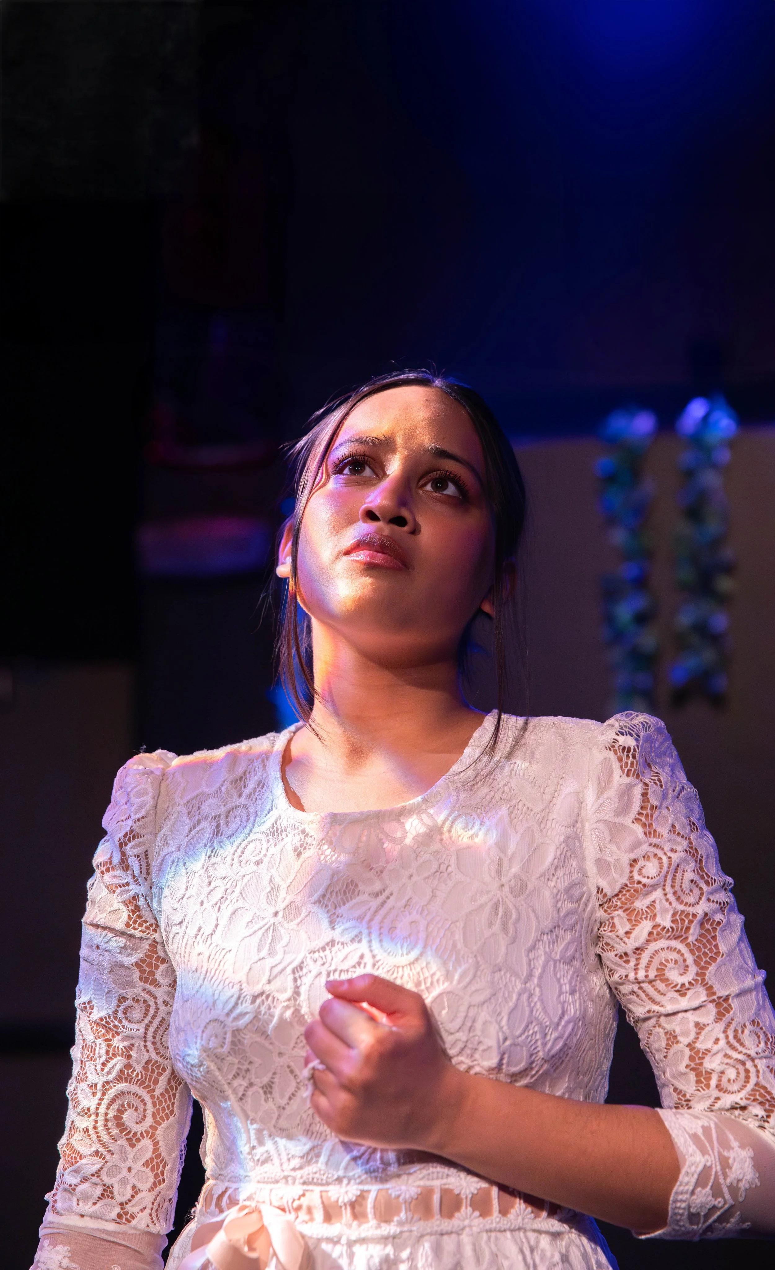 A young woman with dark hair and light skin, wearing a white lace dress, looking thoughtfully into the distance with purple and blue lighting highlighting her face.
