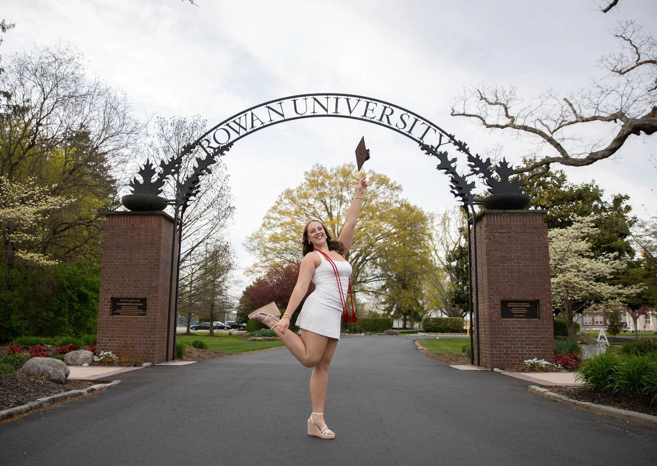 A woman in a white dress and wedge sandals jumping in front of the Iowa University entrance gate, holding a diploma and smiling.