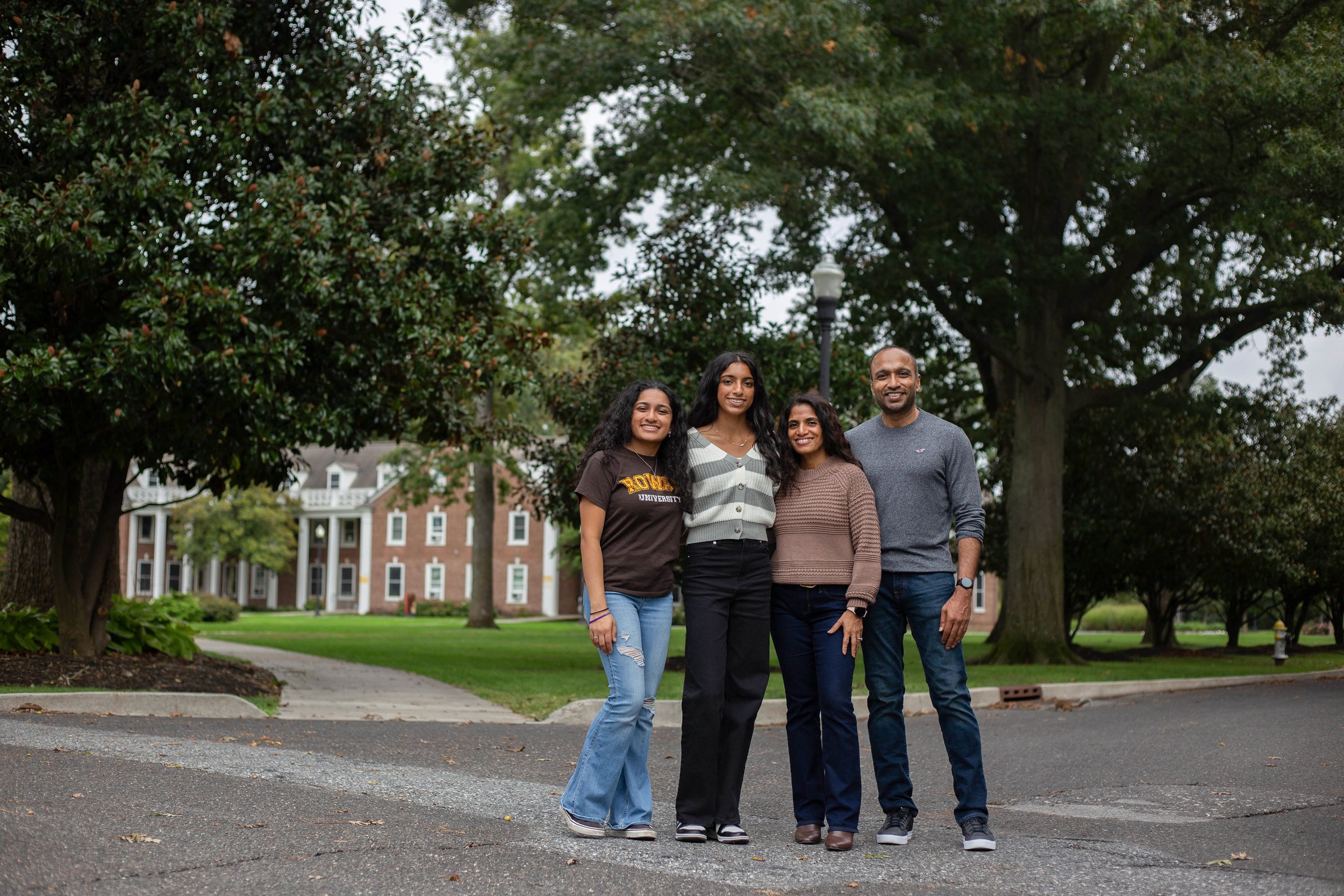 A group of four diverse people, two women and two men, standing together outdoors in front of a large tree and a building on a cloudy day, smiling for the camera.
