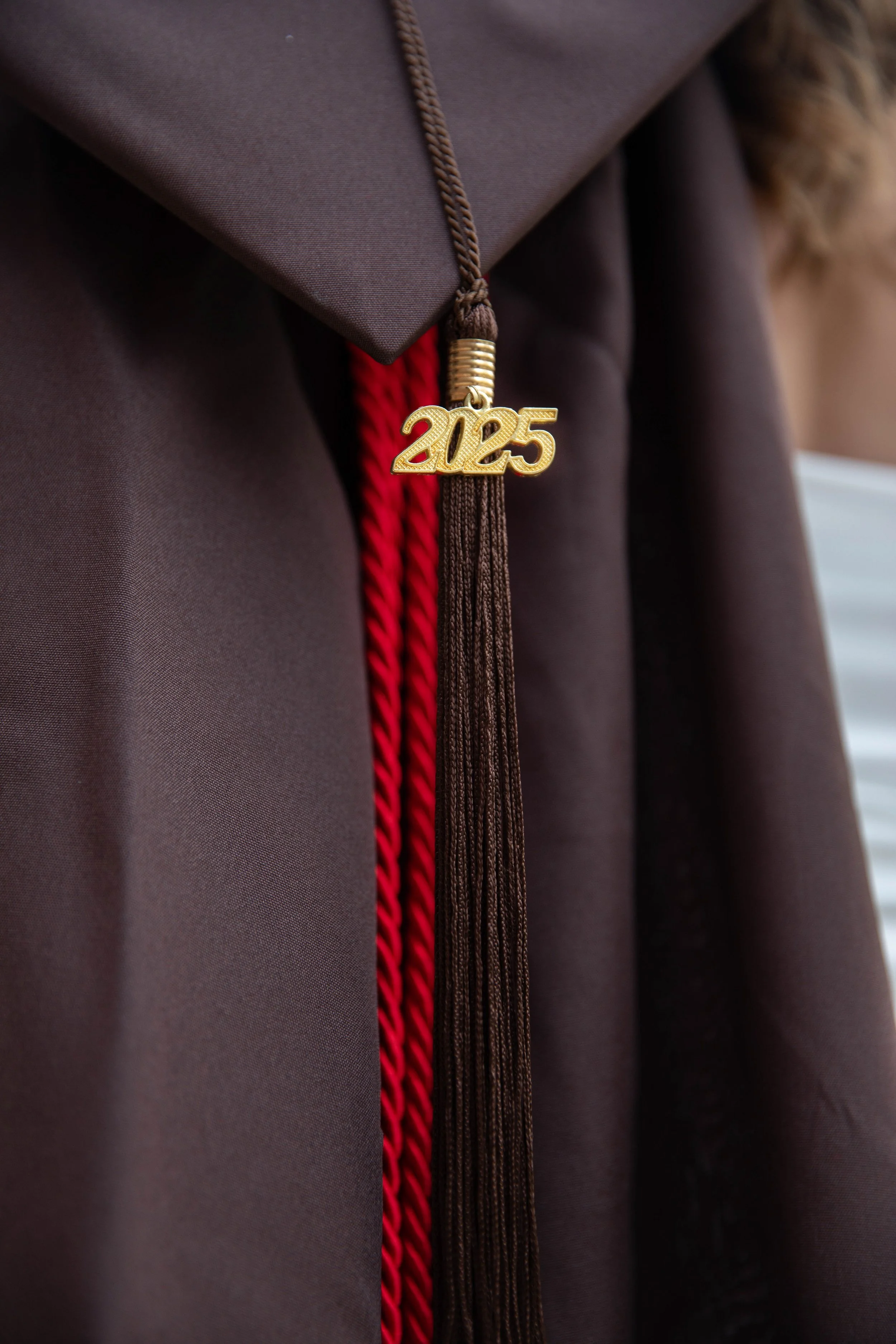 Close-up photo of a graduation cap with a gold '2025' pendant and red and brown cords, worn on a person dressed in a black gown.