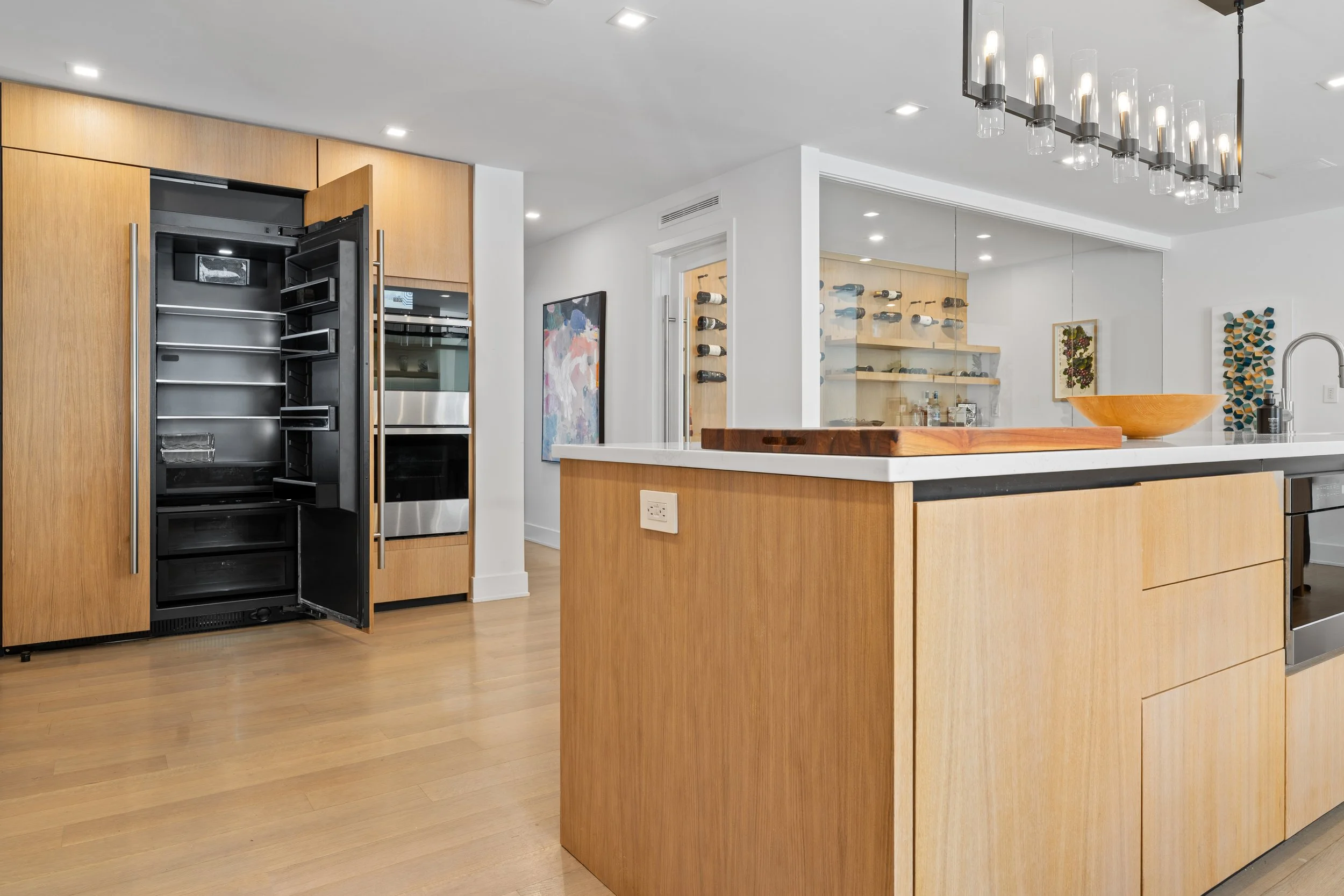 Modern kitchen with wooden cabinets, an open refrigerator, a kitchen island with a wooden bowl, a wine rack, and a ceiling light fixture.