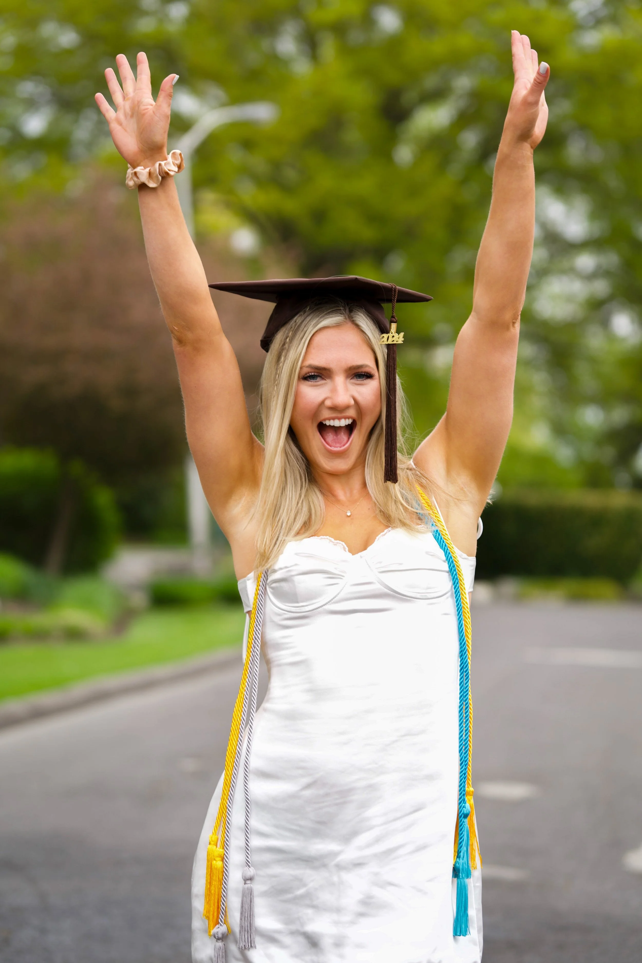 A young woman in a white dress is celebrating her graduation outdoors, wearing a cap with a '2024' tassel, making a joyful expression with arms raised, and wearing multiple graduation cords.