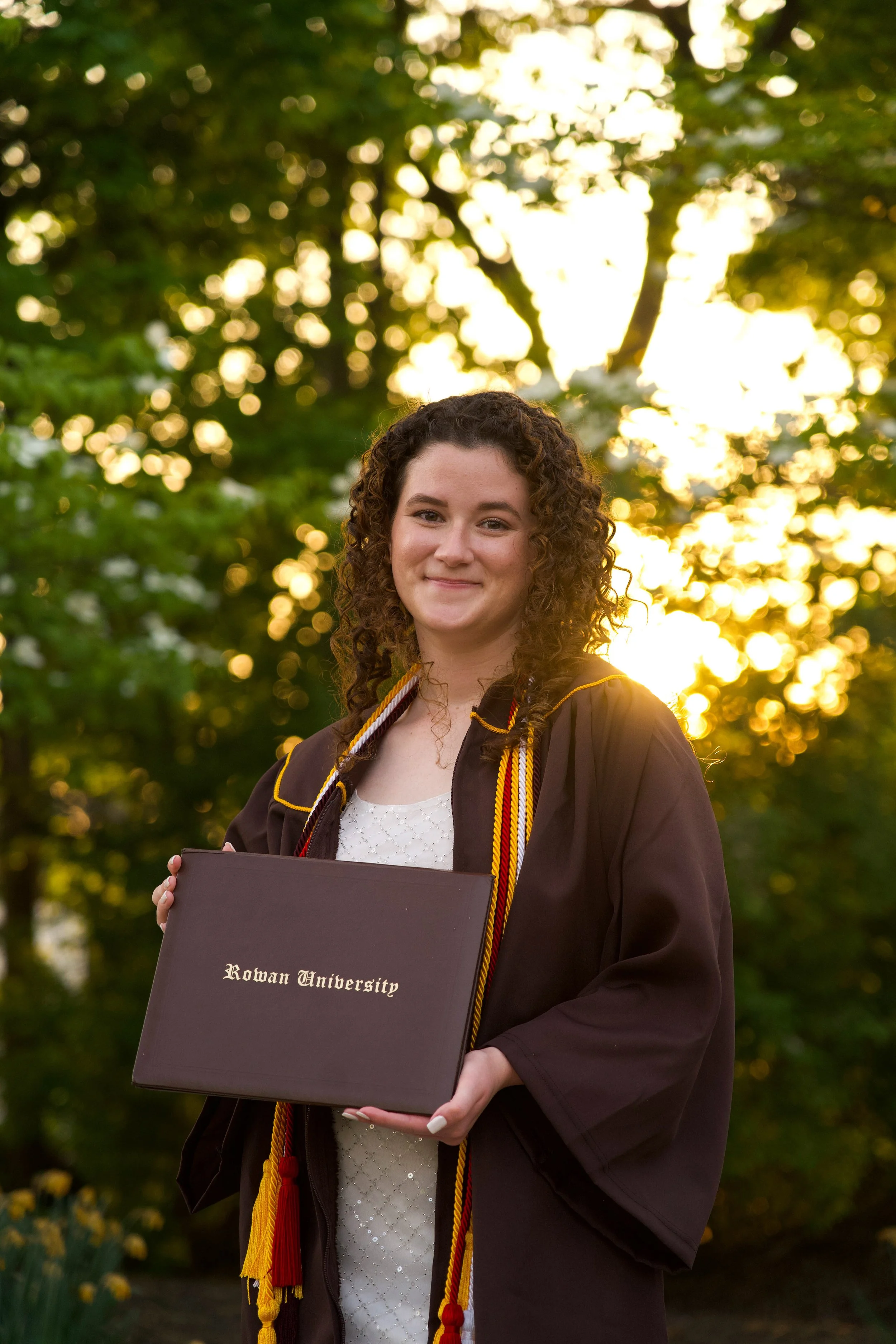 A young woman in graduation gown and cap holding a diploma from Rowan University, standing outdoors in front of green trees with sunlight filtering through its leaves.