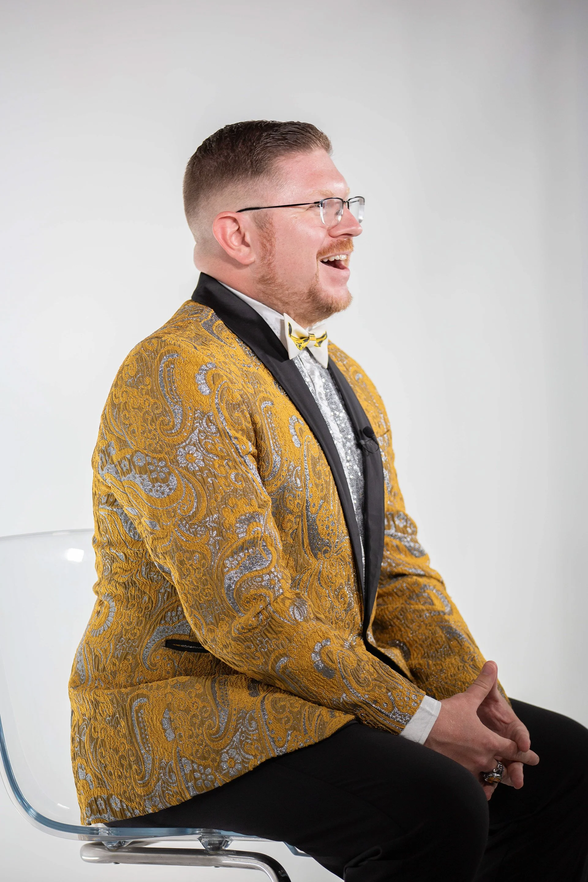 A man with short brown hair, glasses, and a beard, smiling and wearing a yellow and silver patterned tuxedo with a bowtie. He is seated on a transparent chair, against a plain white background.