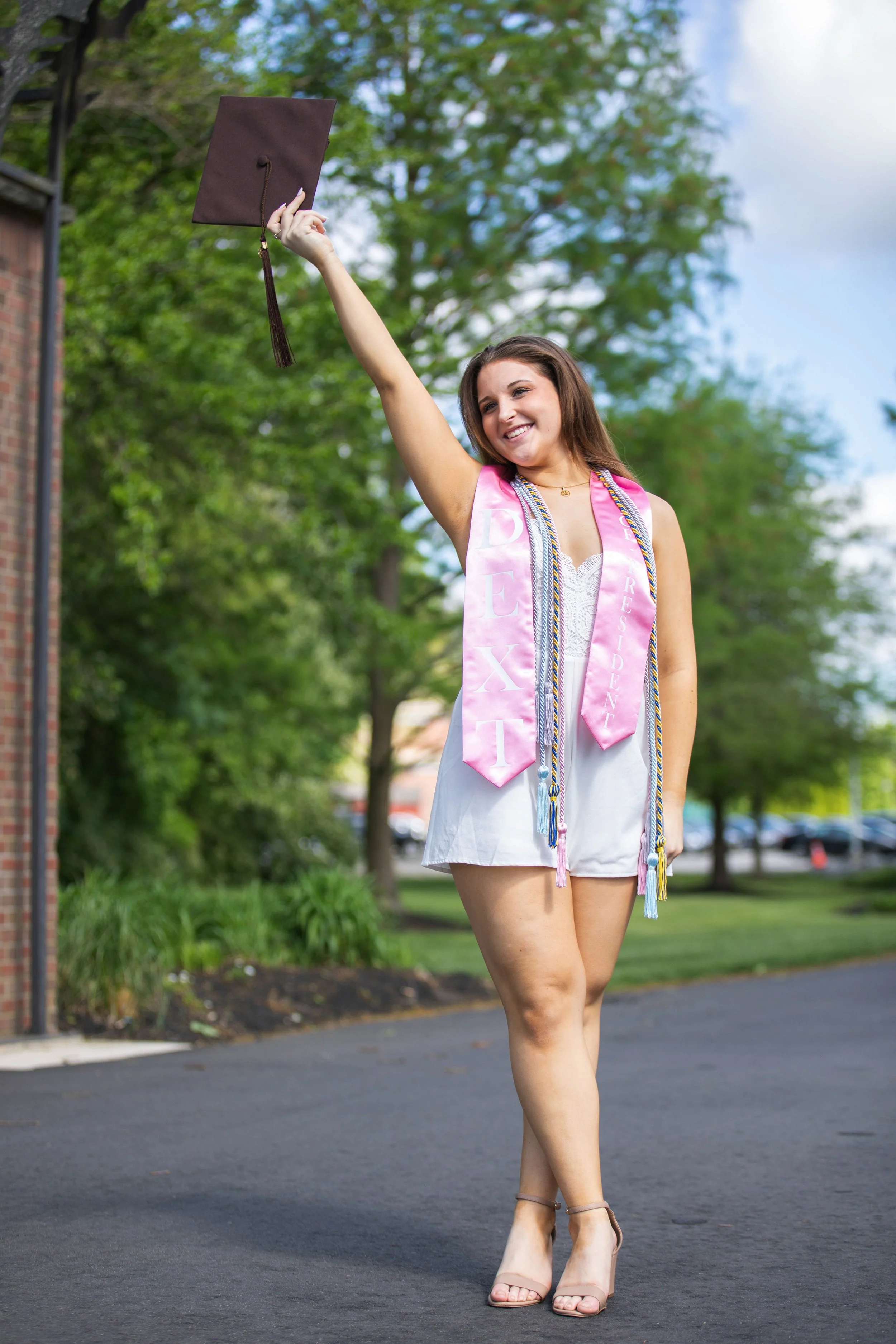 A young woman celebrating her graduation outdoors on a sunny day, wearing a white dress and a pink graduation stole, holding her graduation cap high in the air, smiling, with trees and a brick building in the background.