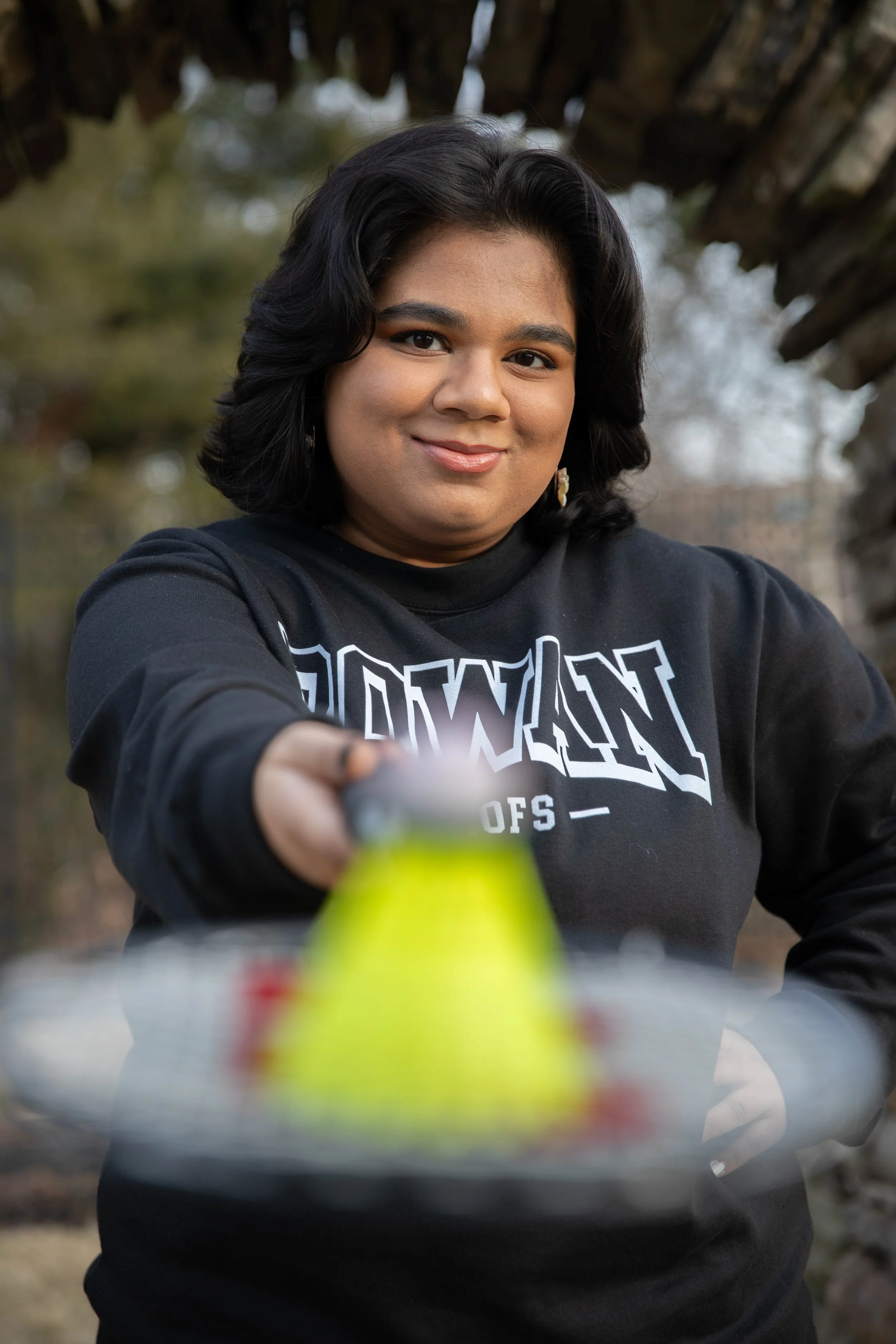 A woman with black hair smiling and taking a selfie outdoors, with trees in the background. She is wearing a black sweatshirt and holding the camera with her right hand.