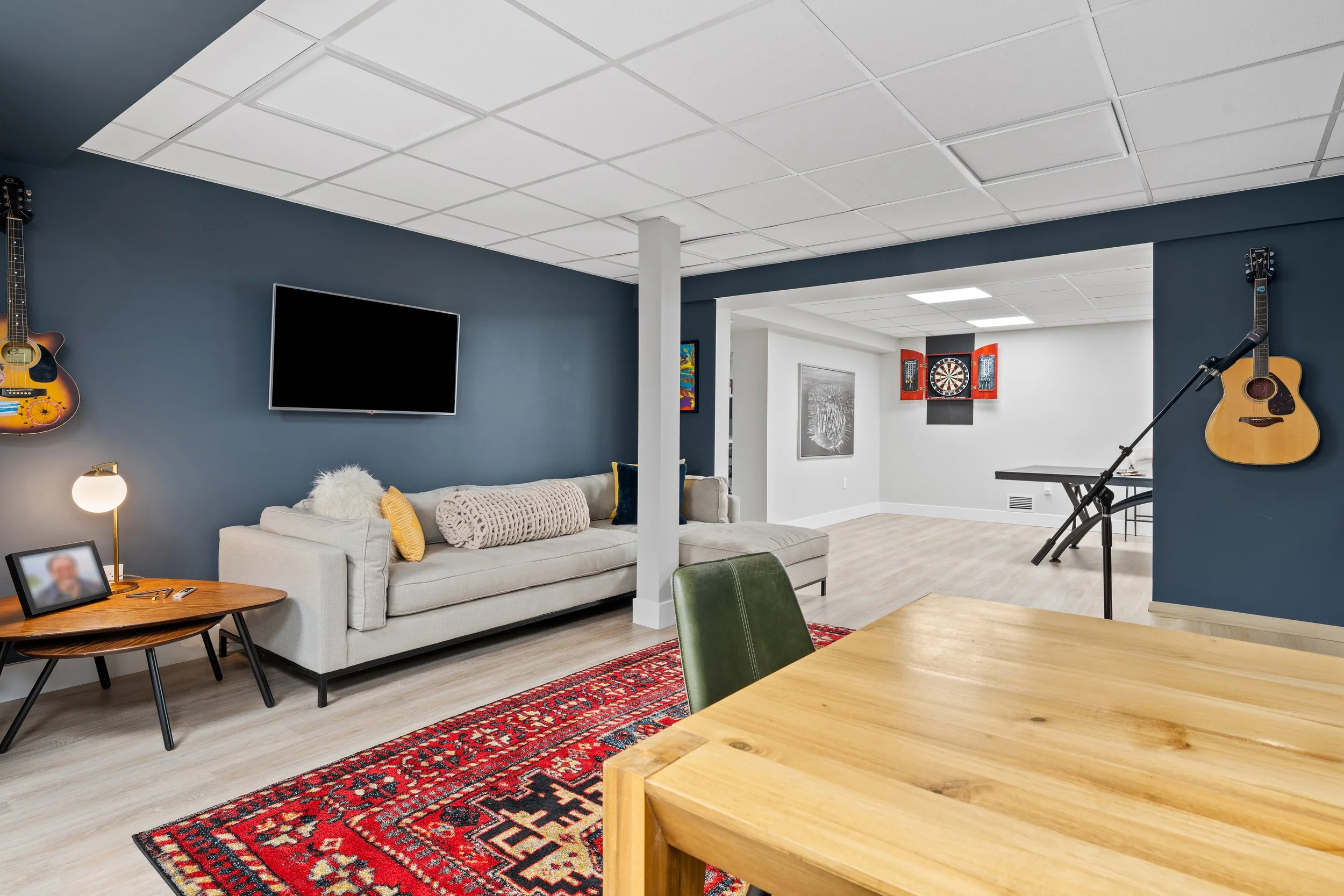 Living room with a gray sofa, a mounted flat-screen TV on a dark blue wall, two acoustic guitars hanging on the wall, a small side table with a framed photo and a lamp, and a red patterned rug. An adjoining space has a dartboard on the wall, musical 