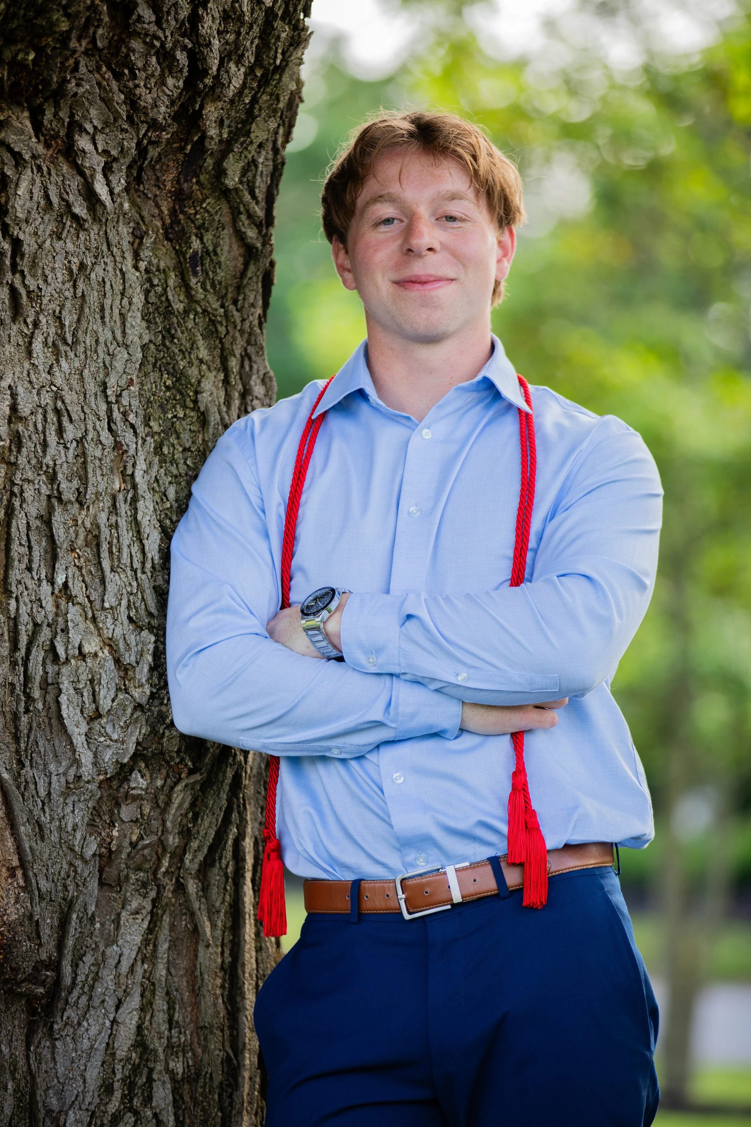A young man with red hair wearing a light blue dress shirt, dark blue pants, and a brown belt, standing outdoors next to a large tree with green blurred foliage in the background. He has a red cord around his neck and is smiling with arms crossed.