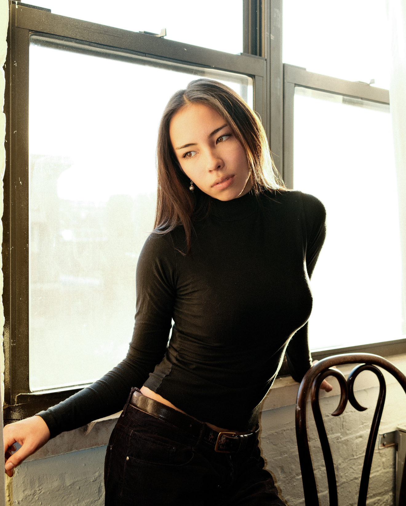 A young woman with long dark hair wearing a black turtleneck sweater and black pants, leaning against a window with sunlight shining through behind her.