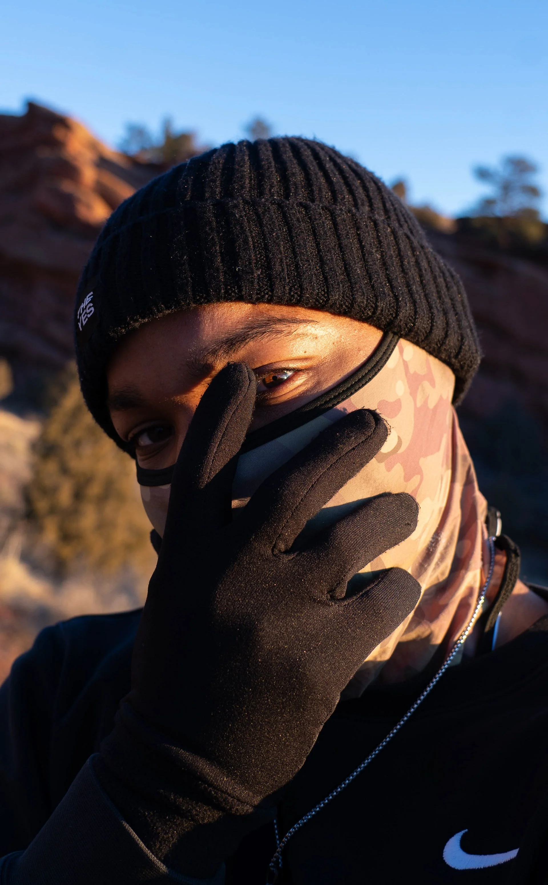 A person wearing a black beanie, a camouflage-patterned face covering, black gloves, and a black jacket with a Nike logo, making a peace sign near their eye outdoors during sunset or sunrise.