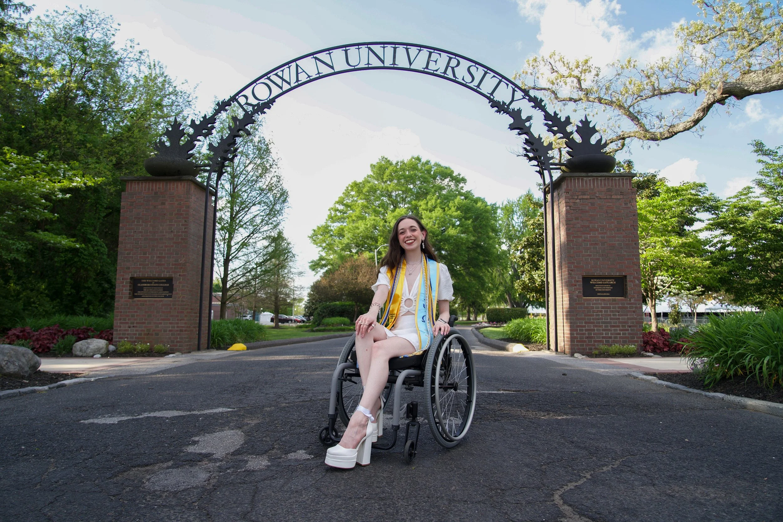 A woman in a wheelchair smiling at the camera stands in front of the entrance to a university, with trees and a brick gate archway that reads 'Browan University' visible in the background.