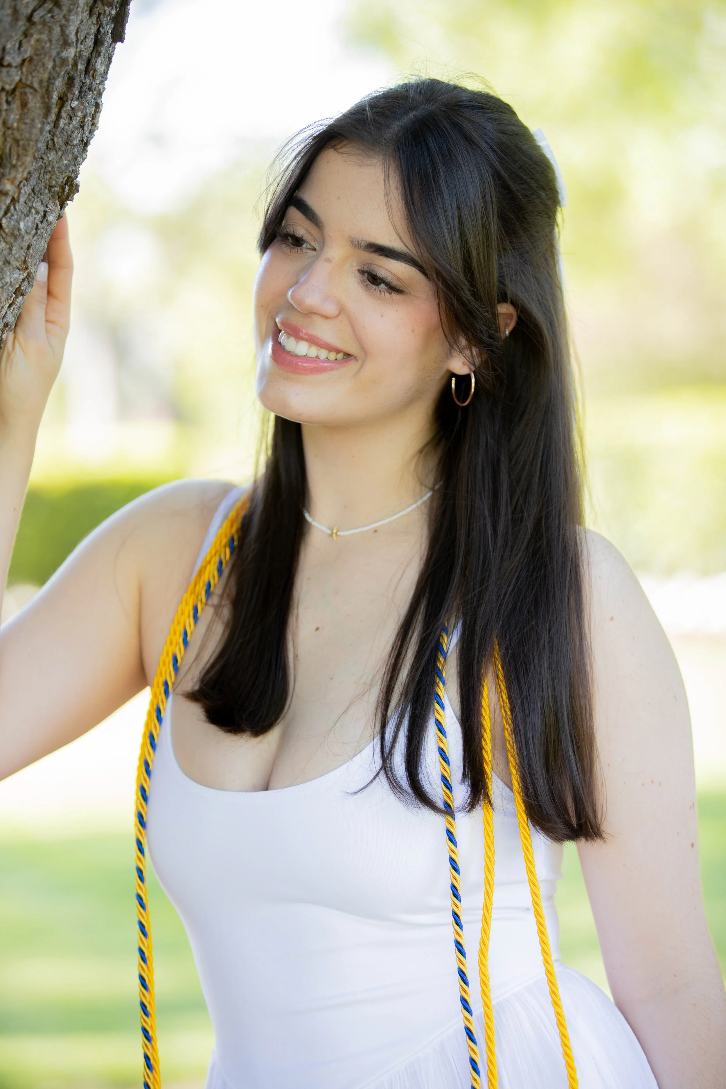 A young woman with long dark hair, wearing a white dress, is outdoors, smiling, and touching a tree with her left hand. She has gold hoop earrings, a choker necklace, and yellow, blue, and black cords hanging from her shoulders, possibly part of a gr