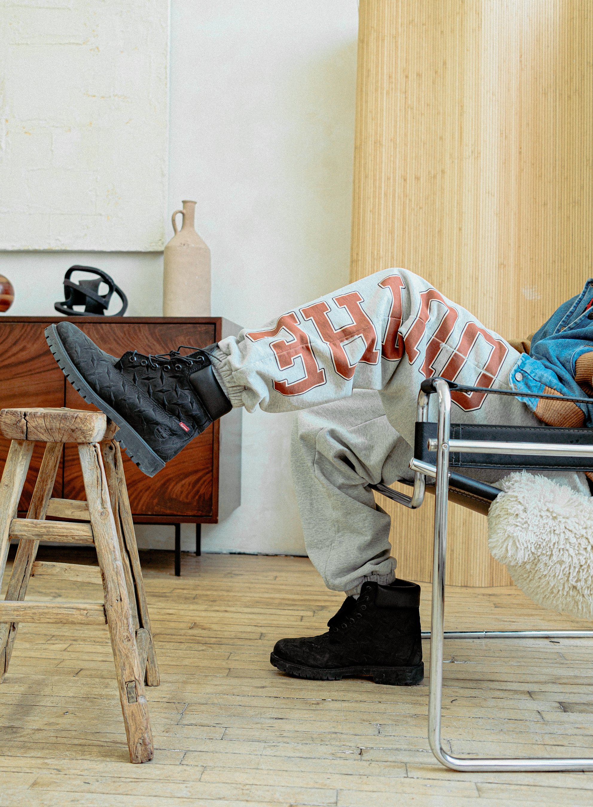 Person sitting in a wheelchair wearing black boots, gray sweatpants with red lettering, and a blue jacket, with their legs crossed and feet resting on a wooden stool, in a room with wooden flooring and a wooden wall.