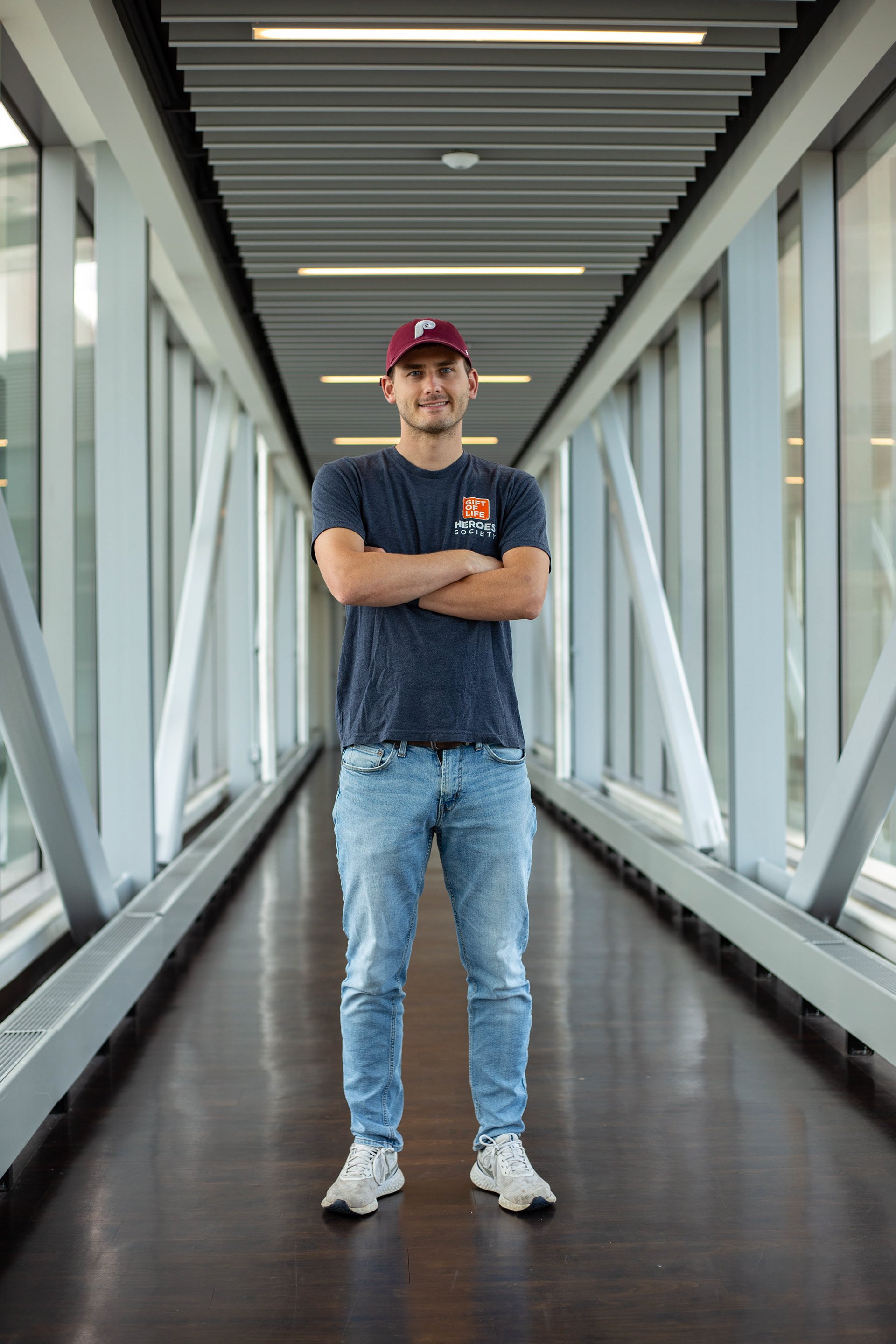 A young man standing in a modern, glass-enclosed walkway with wooden flooring, smiling with arms crossed, wearing a maroon cap, a dark gray T-shirt with logos, jeans, and white sneakers.