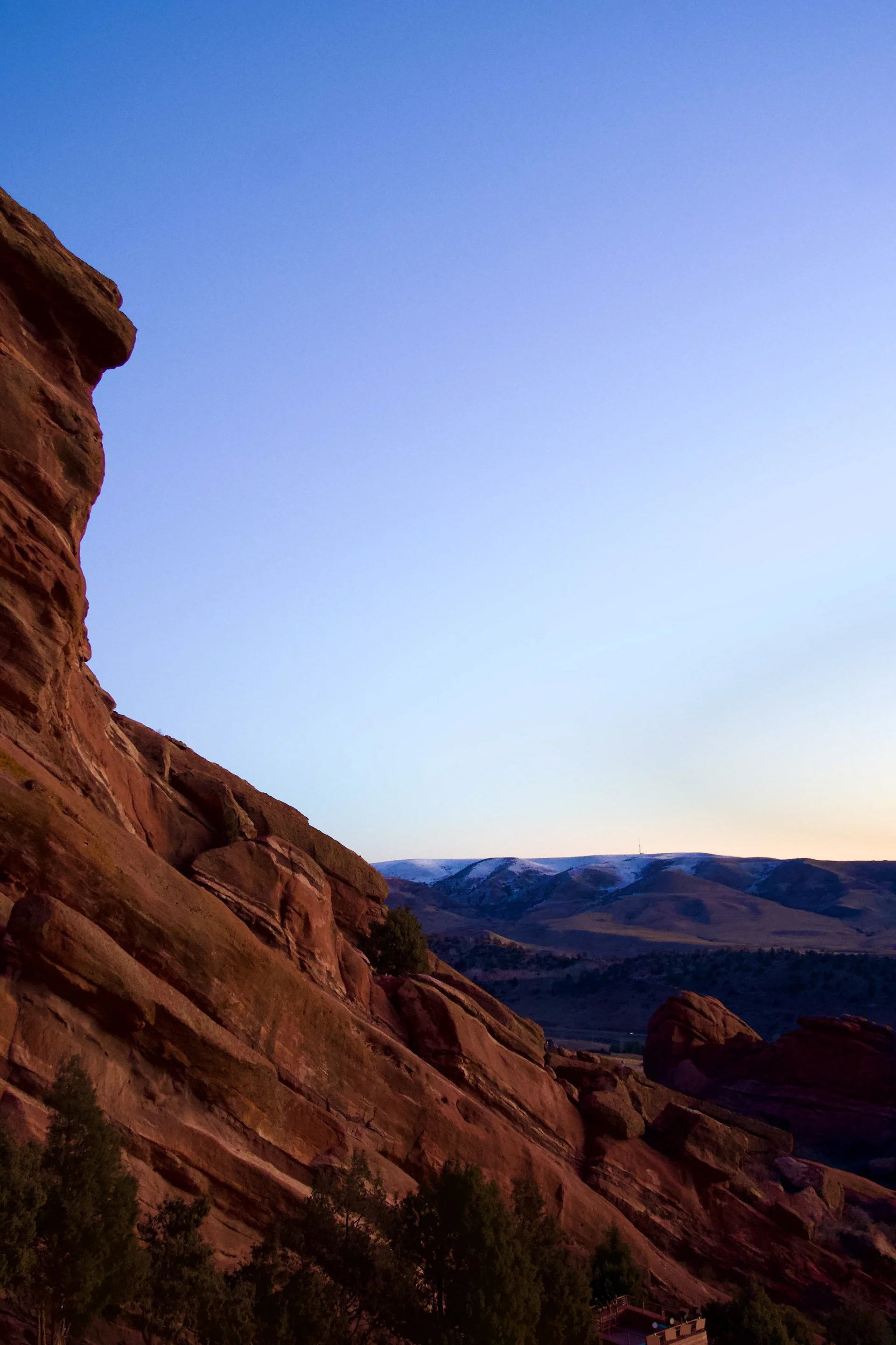 A rugged, reddish-brown mountain with a steep, layered surface on the left side, and a series of rolling, dark blue and purple distant mountain ridges with snow-capped peaks in the background under a clear blue sky at dusk.