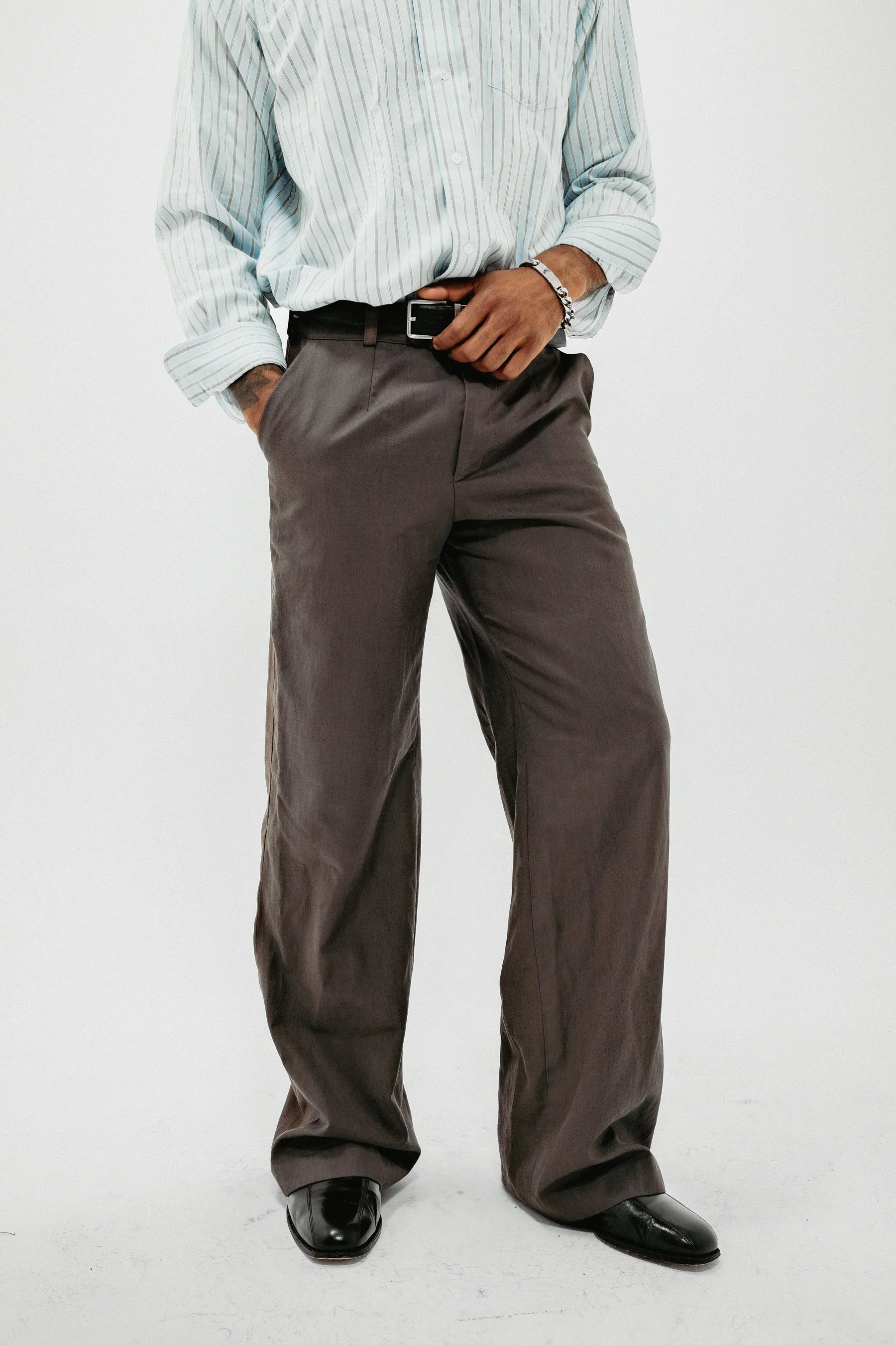 Person wearing a light blue and white striped shirt, gray trousers, black shoes, and a bracelet, standing against a plain white background.
