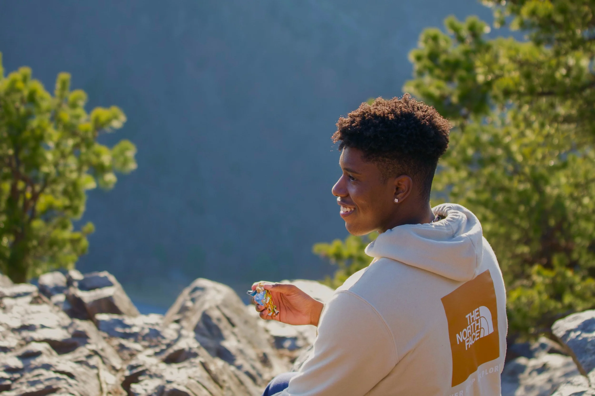 Young woman with short curly hair smiling outdoors, holding a snack, wearing a hoodie with The North Face logo, surrounded by greenery and rocks.