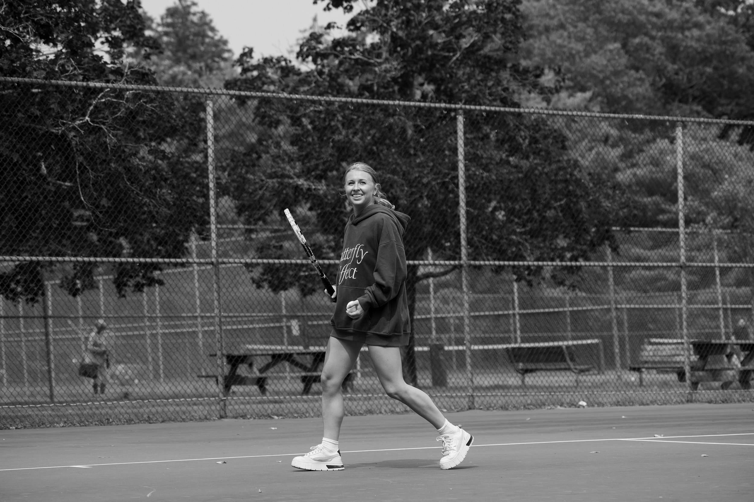 A young woman playing tennis on an outdoor court, smiling, wearing a hoodie with the words 'Butterfly Effect,' running with a tennis racket in her right hand, in front of a chain-link fence and trees.