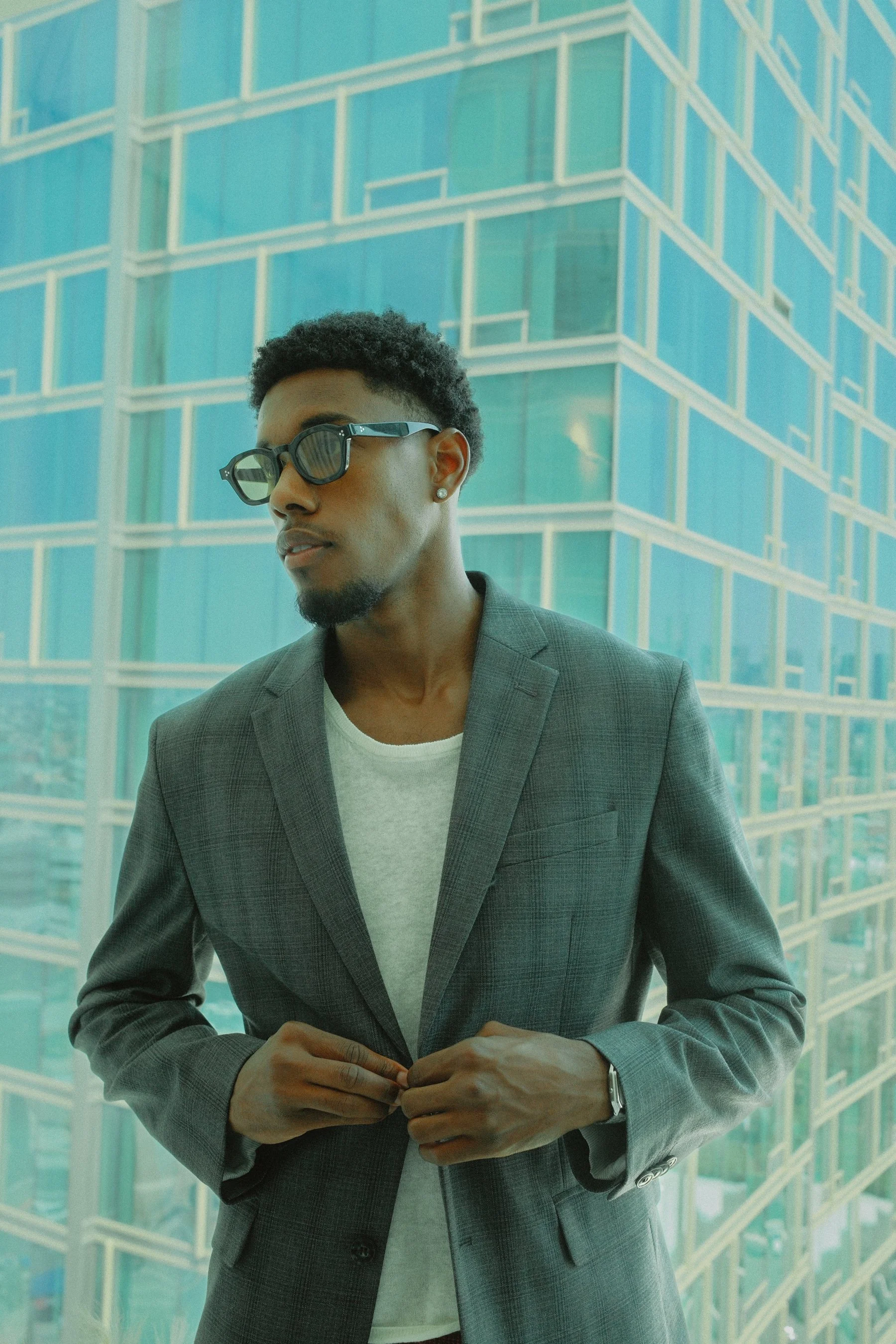 A man wearing sunglasses, a gray blazer, and a white t-shirt is standing in front of a modern glass building with a geometric pattern.