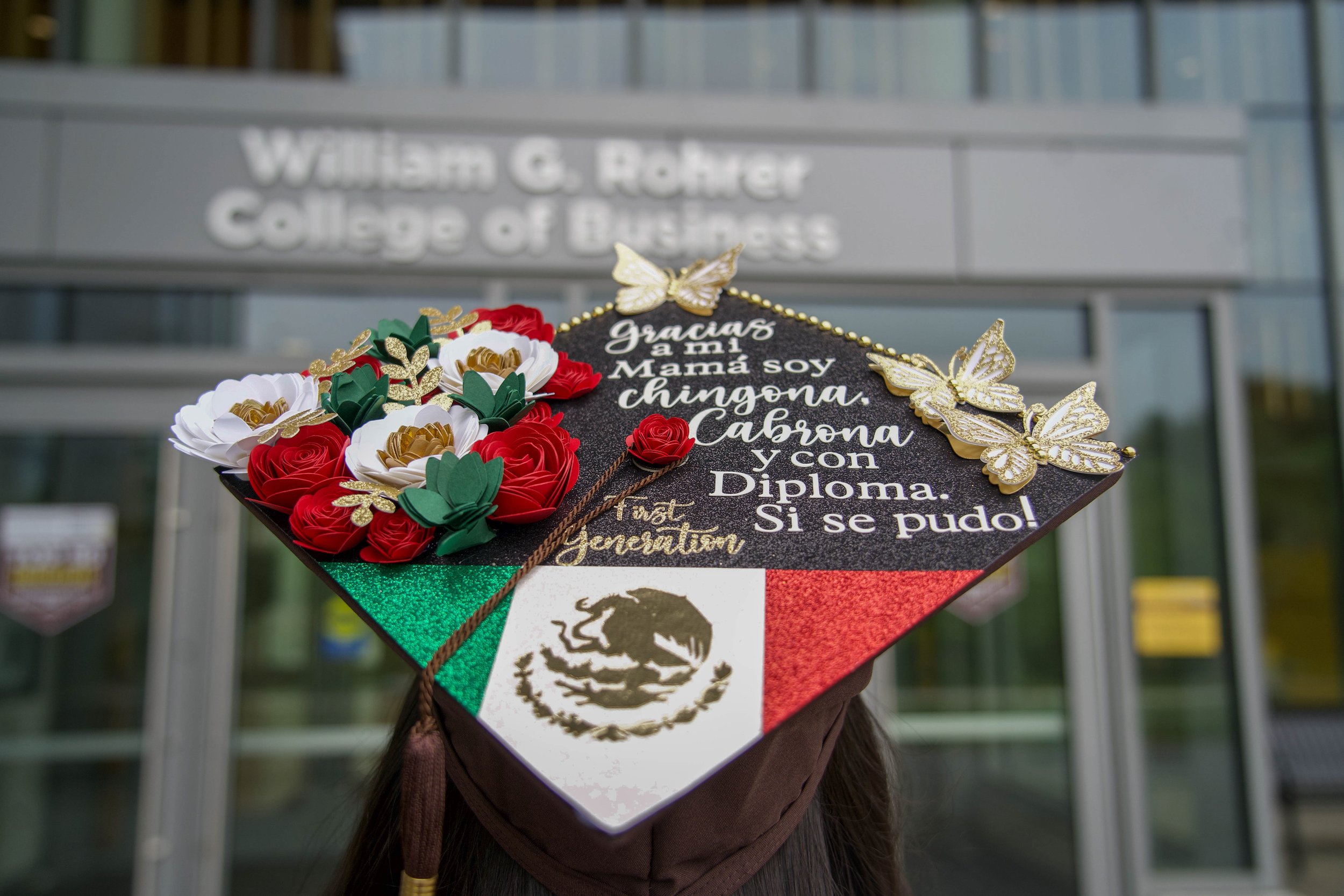 A graduation cap decorated with flowers, gold butterflies, and Mexican flag colors, in front of a William G. Basler College of Business sign.