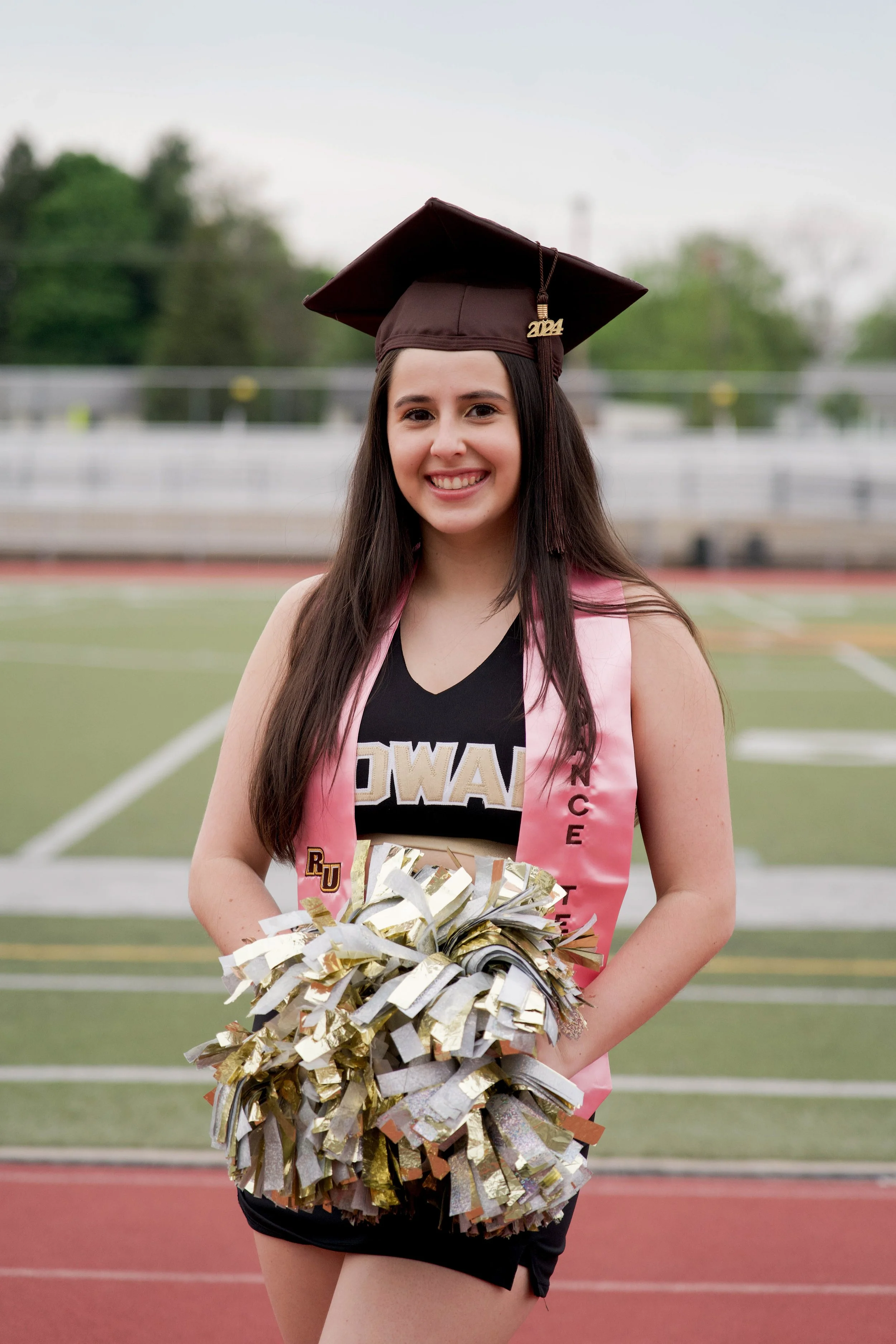 A young woman with long dark hair dressed in a cheerleader outfit standing on a track field. She is wearing a black sports bra, black shorts, a pink sash, and a maroon graduation cap with a '2024' tassel. She is smiling, holding gold and silver pom-p