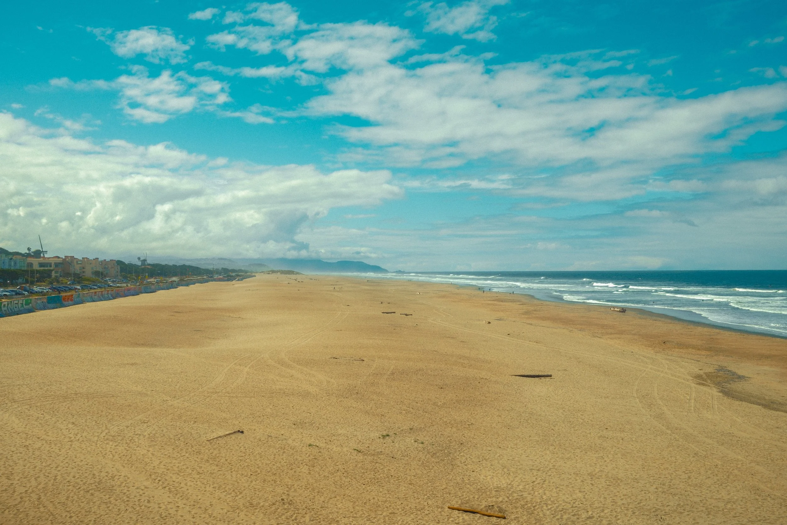 A sandy beach with a clear blue sky, scattered clouds, ocean waves, and distant houses along the shoreline.