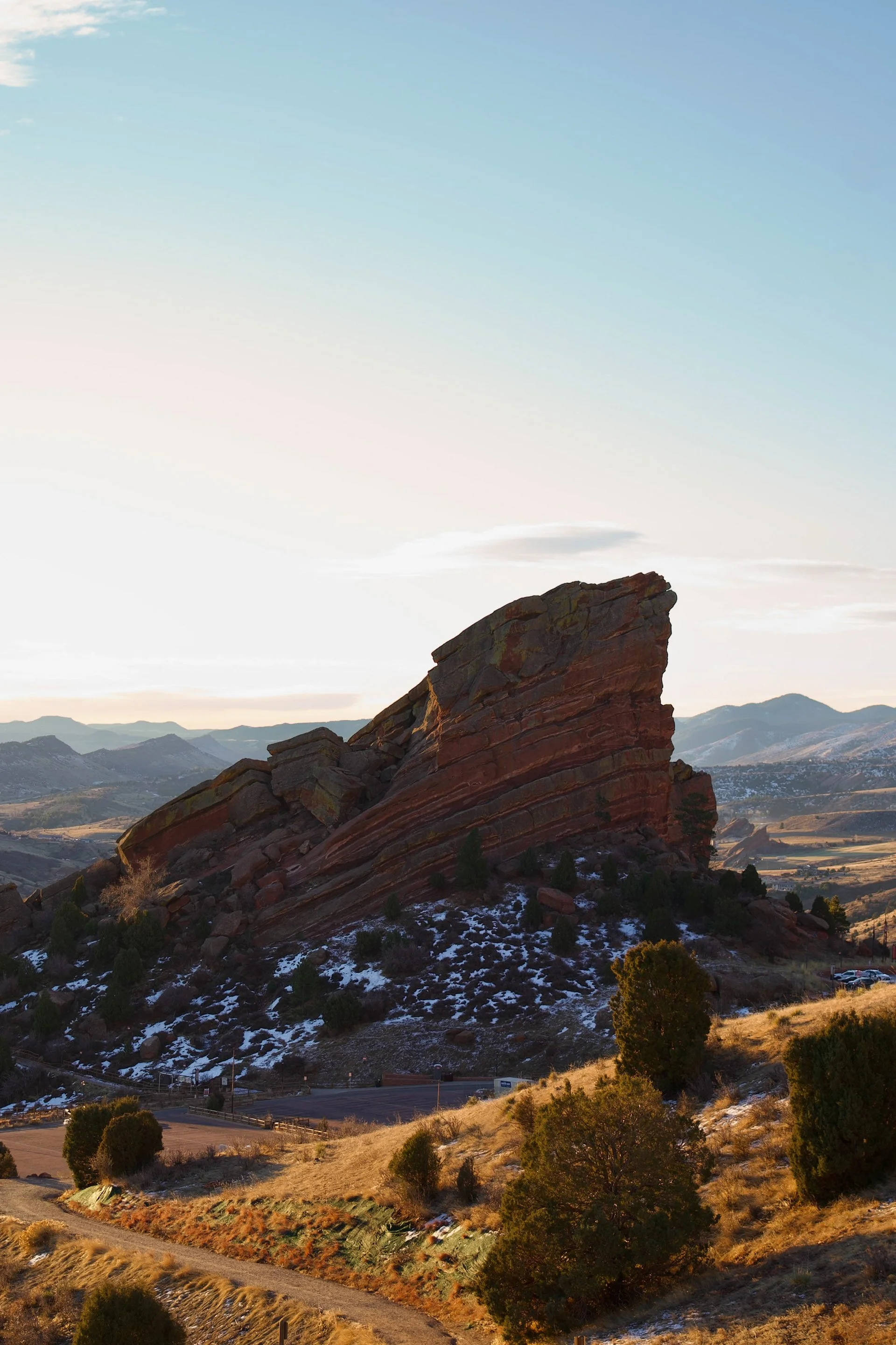 Large reddish rock formation and hills with patches of snow, trees, and a road in the foreground, mountains in the distance under a partly cloudy sky.