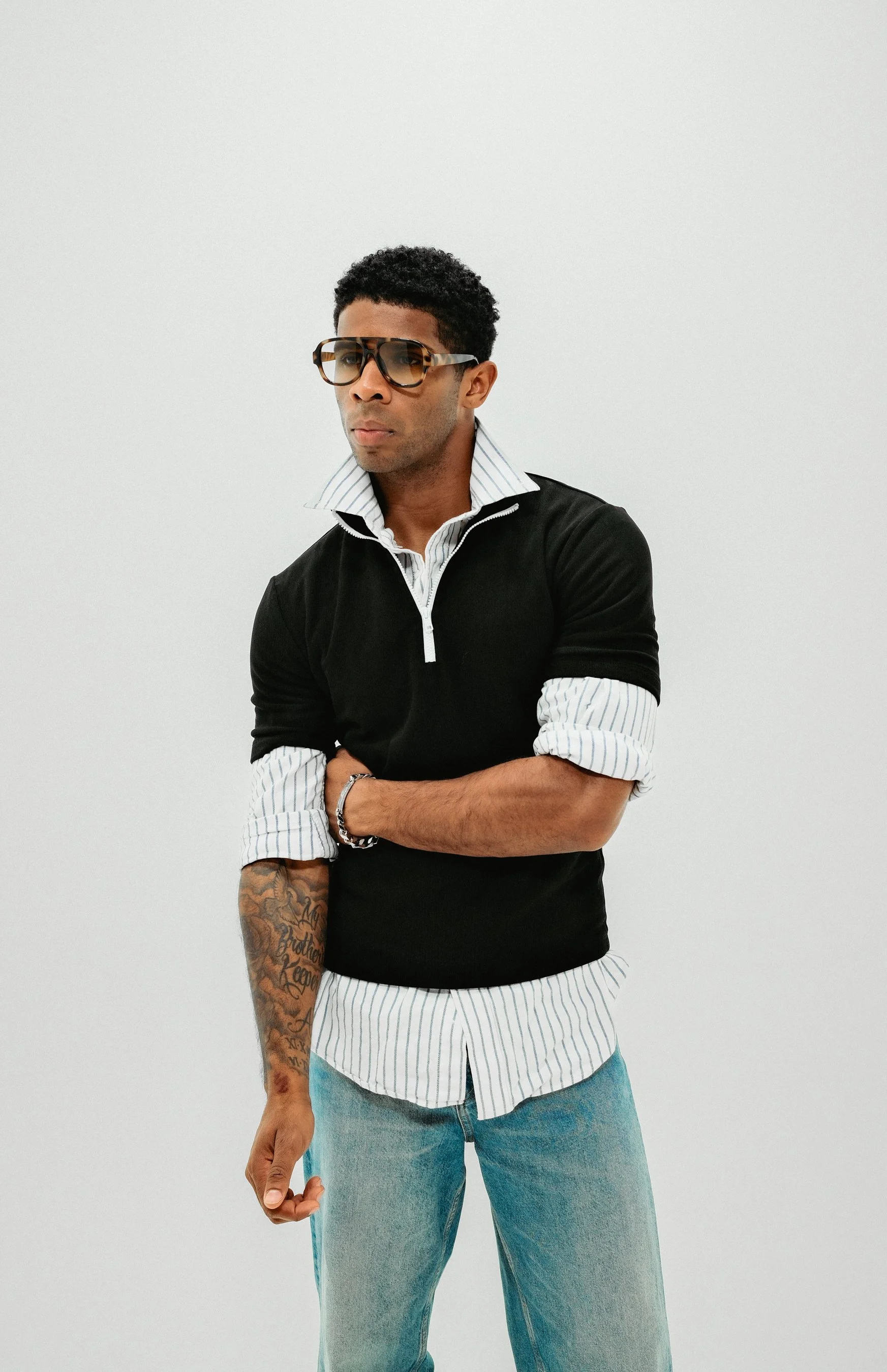A stylish man with curly hair and sunglasses, wearing a layered outfit with a black quarter-zip sweater over a white and blue striped shirt, paired with light blue jeans, standing against a plain white background.