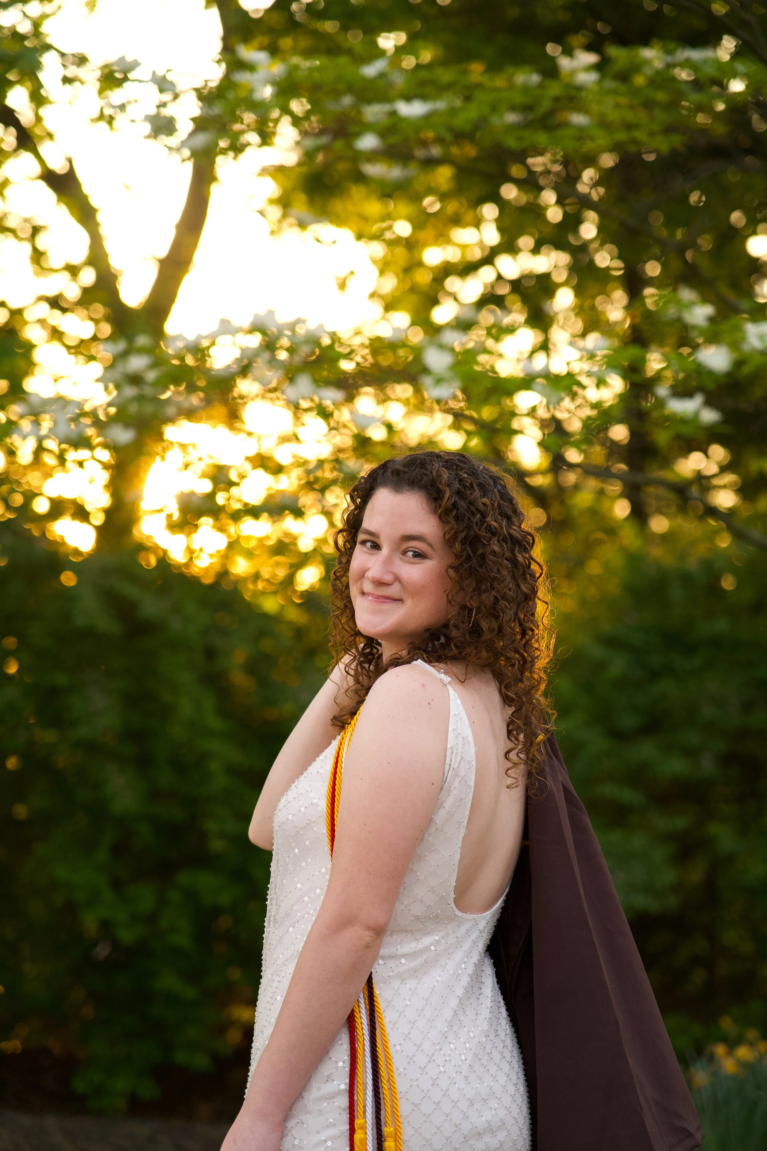 A young woman with curly brown hair is smiling outdoors during sunset, wearing a white sleeveless dress with small embellishments and a graduation stole, with a brown graduation gown draped over her arm, and trees in the background with golden sunlig