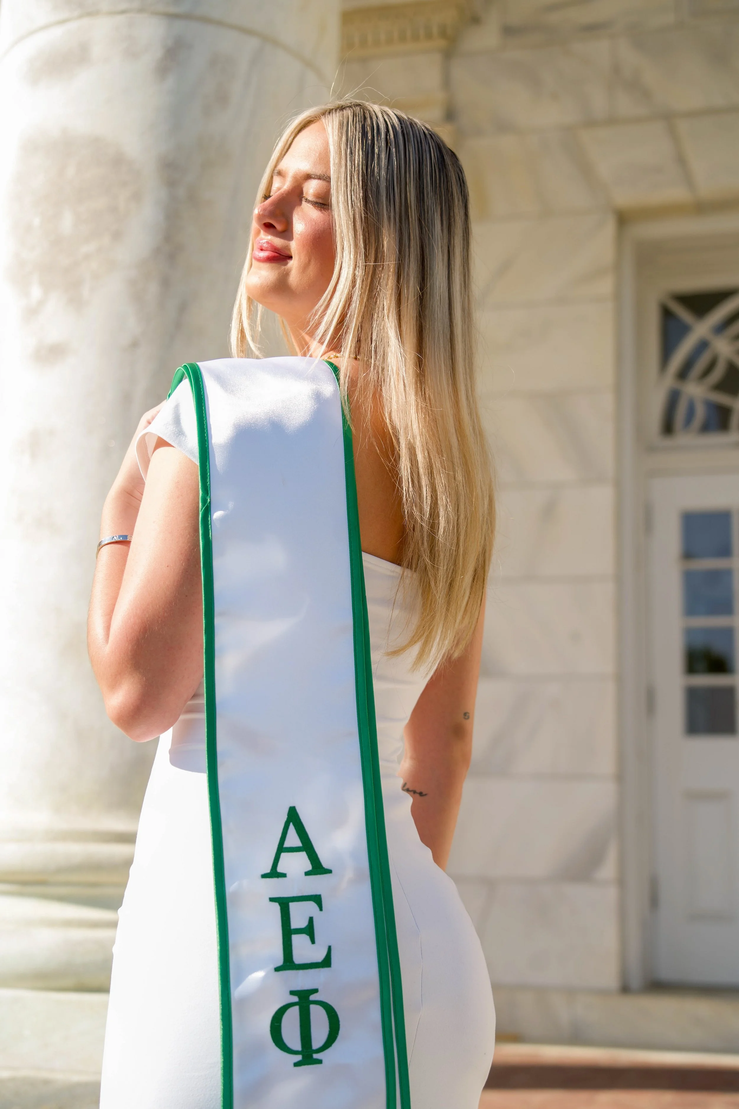 A woman with blonde hair and a white dress standing outside of a stone building with a large window, holding a white bag with green trim and Greek letters.