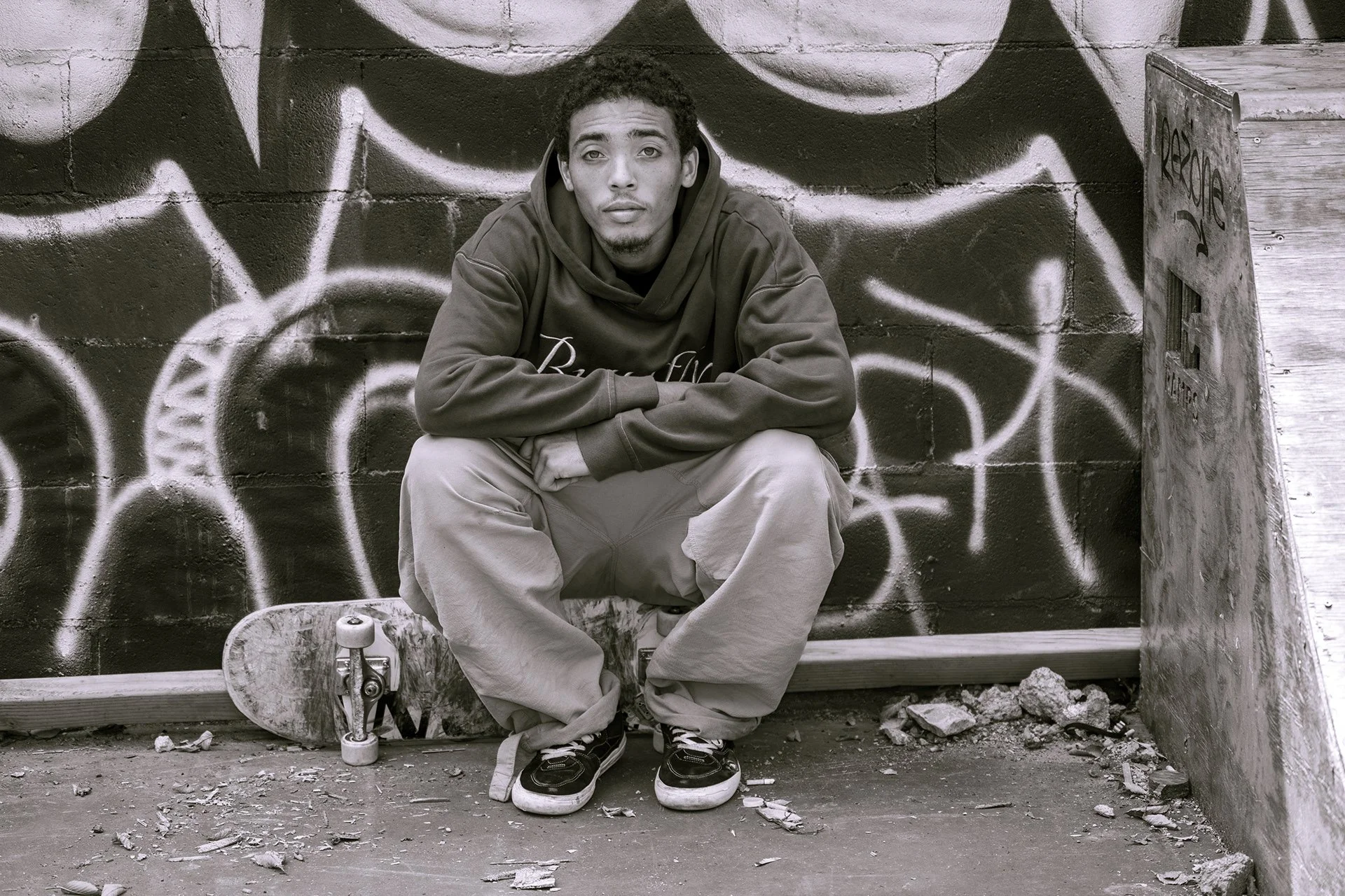A young man with curly hair sitting on a skateboard in front of a graffiti-covered wall, wearing a hoodie and baggy pants, with a skateboard in front of him and debris on the ground.