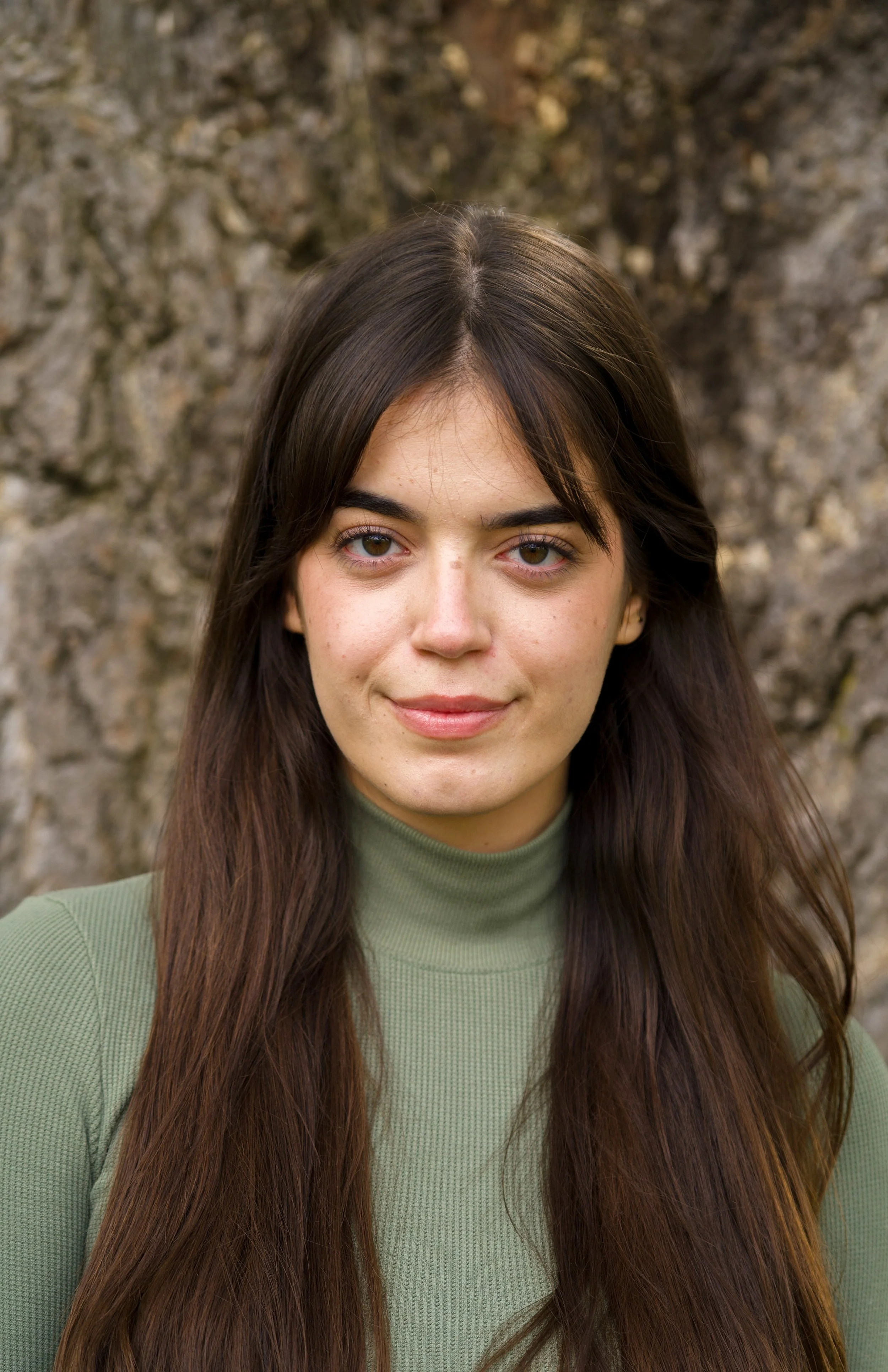 A woman with long dark hair and hazel eyes standing outdoors in front of a textured stone wall, wearing a green long-sleeve top.