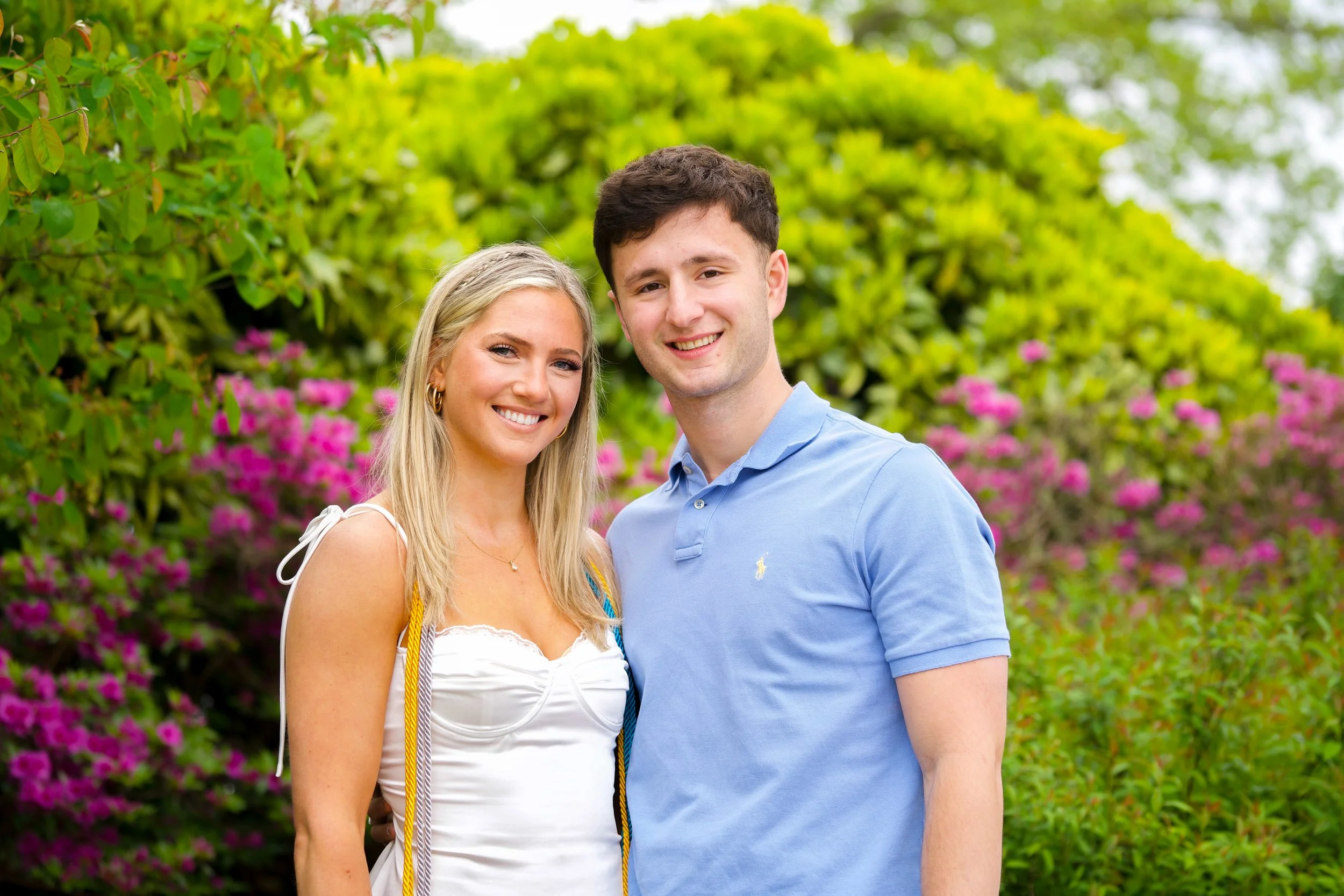 A young woman and a young man smiling and standing close together in front of green foliage and pink flowers.