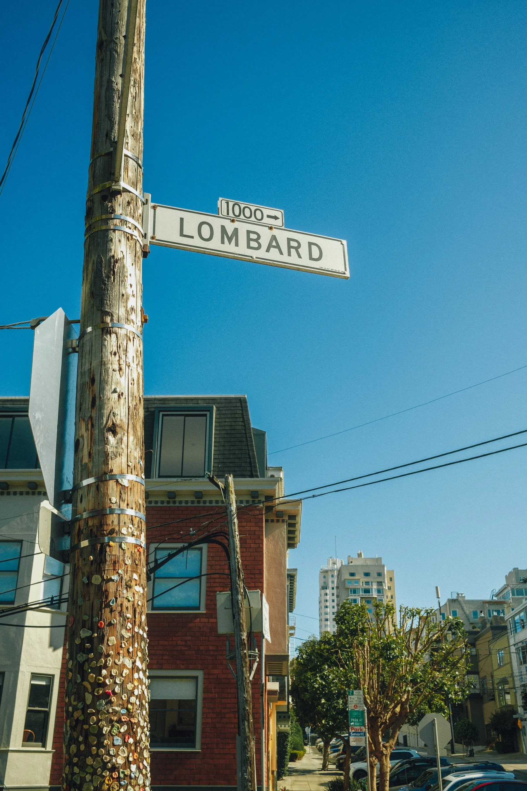 Street view with attached street name sign 'Lombard', utility pole with peeling paint and stickers, residential buildings, trees lining the sidewalk, and blue sky.
