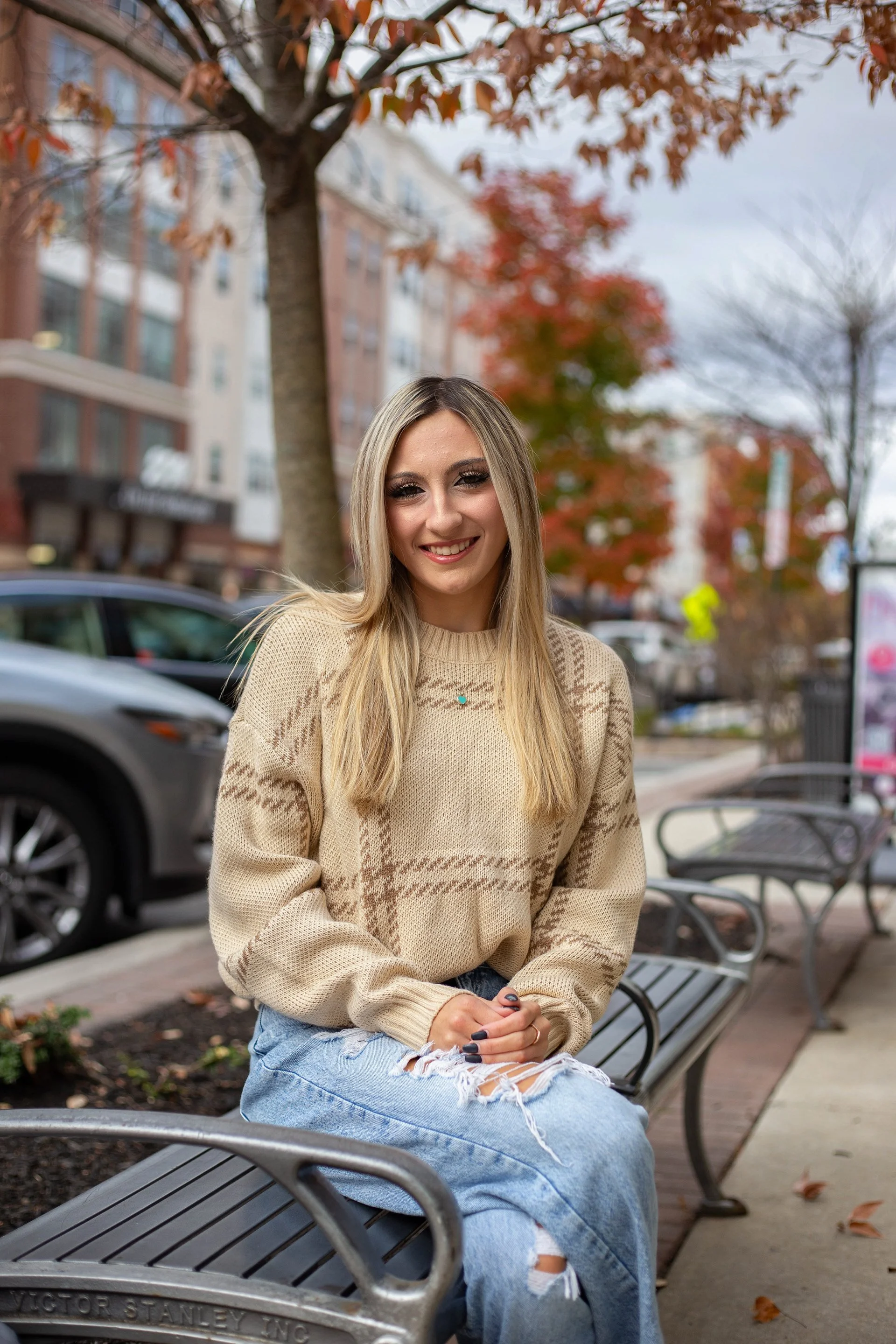 A young woman with long blonde hair sitting on a public park bench during autumn, smiling at the camera, wearing a beige sweater and ripped jeans, with trees showing fall foliage in the background.