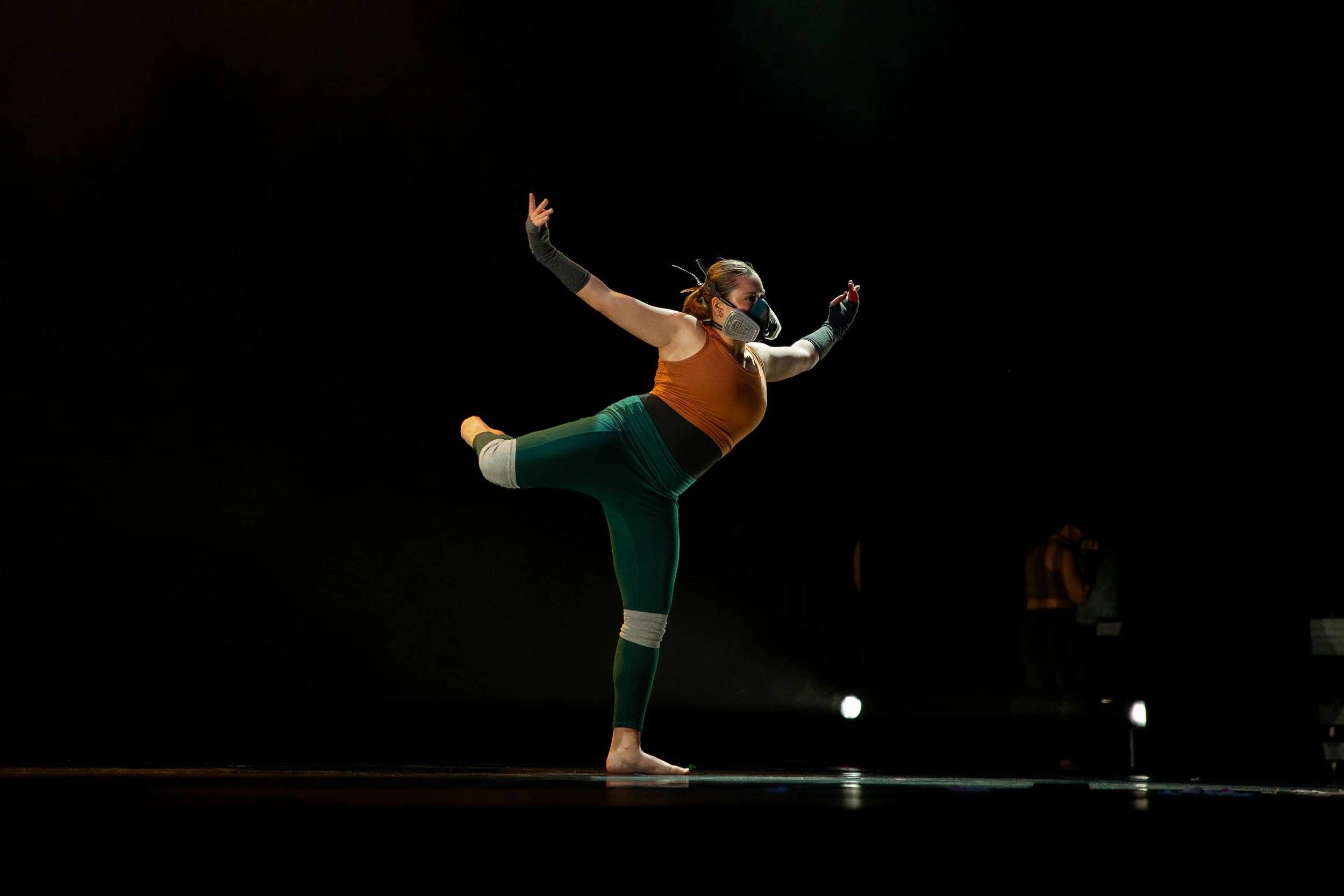 A woman performing a yoga pose on stage, balancing on one leg with the other leg extended back, arms raised, wearing workout clothing, a face mask, and protective arm gear.