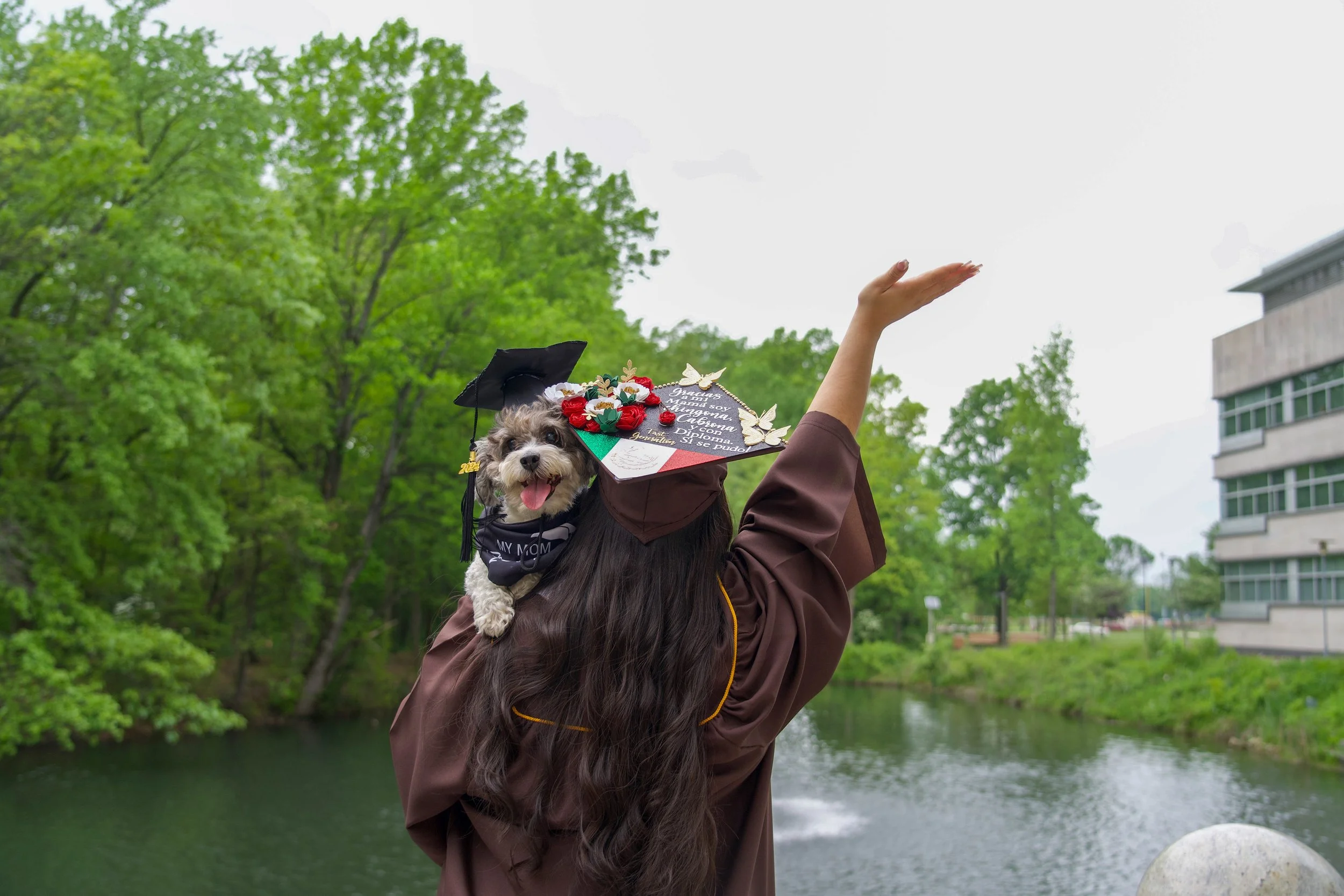A person wearing a brown graduation gown and black cap holds a small dog with a graduation cap and a decorated diploma in front of green trees and a river in an outdoor setting.