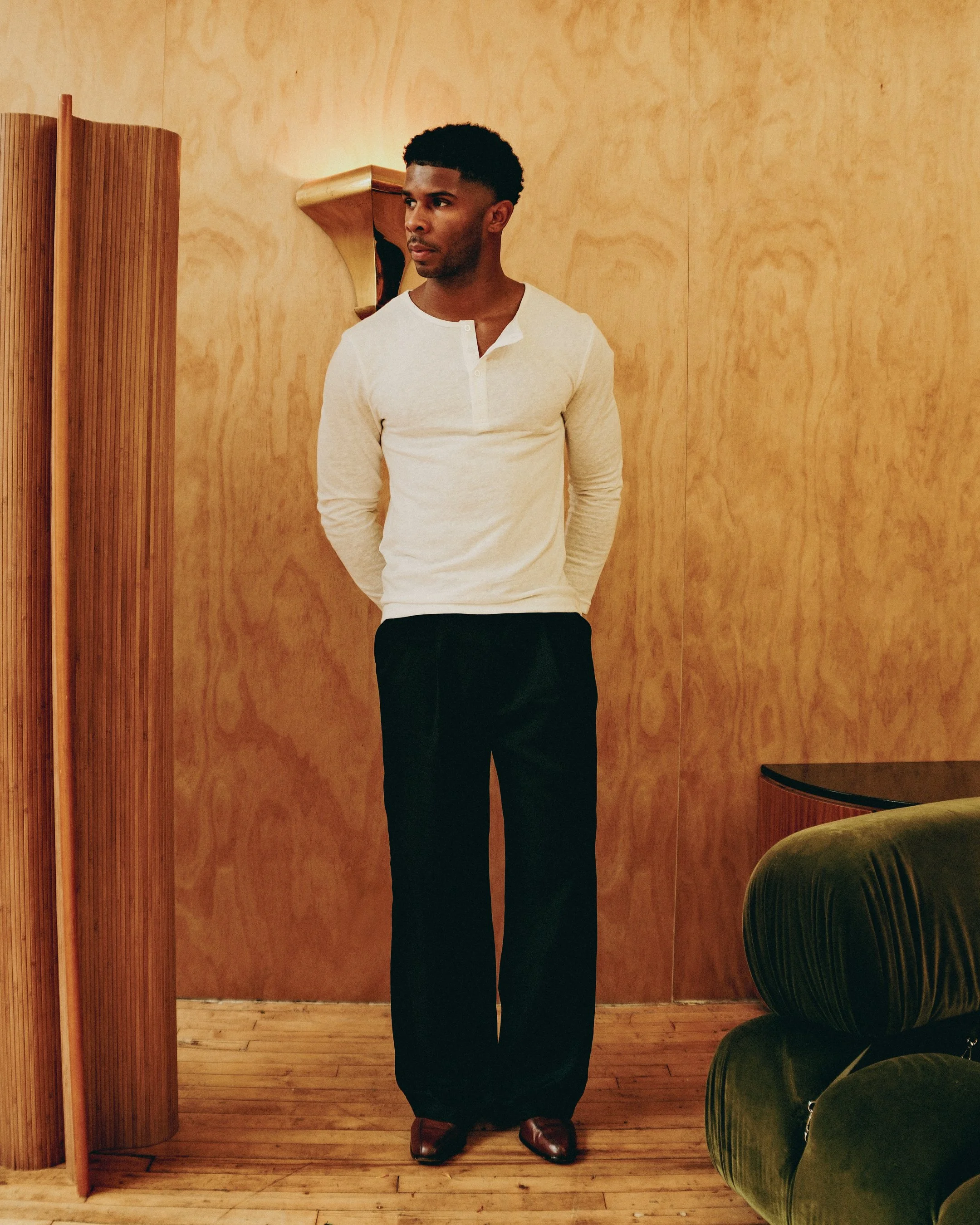 A man stands in a room with wooden walls and floors, dressed in a light-colored long-sleeve shirt, black pants, and brown shoes. He has short, curly black hair and is looking to his left, with his arms behind his back, next to a small green velvet ch