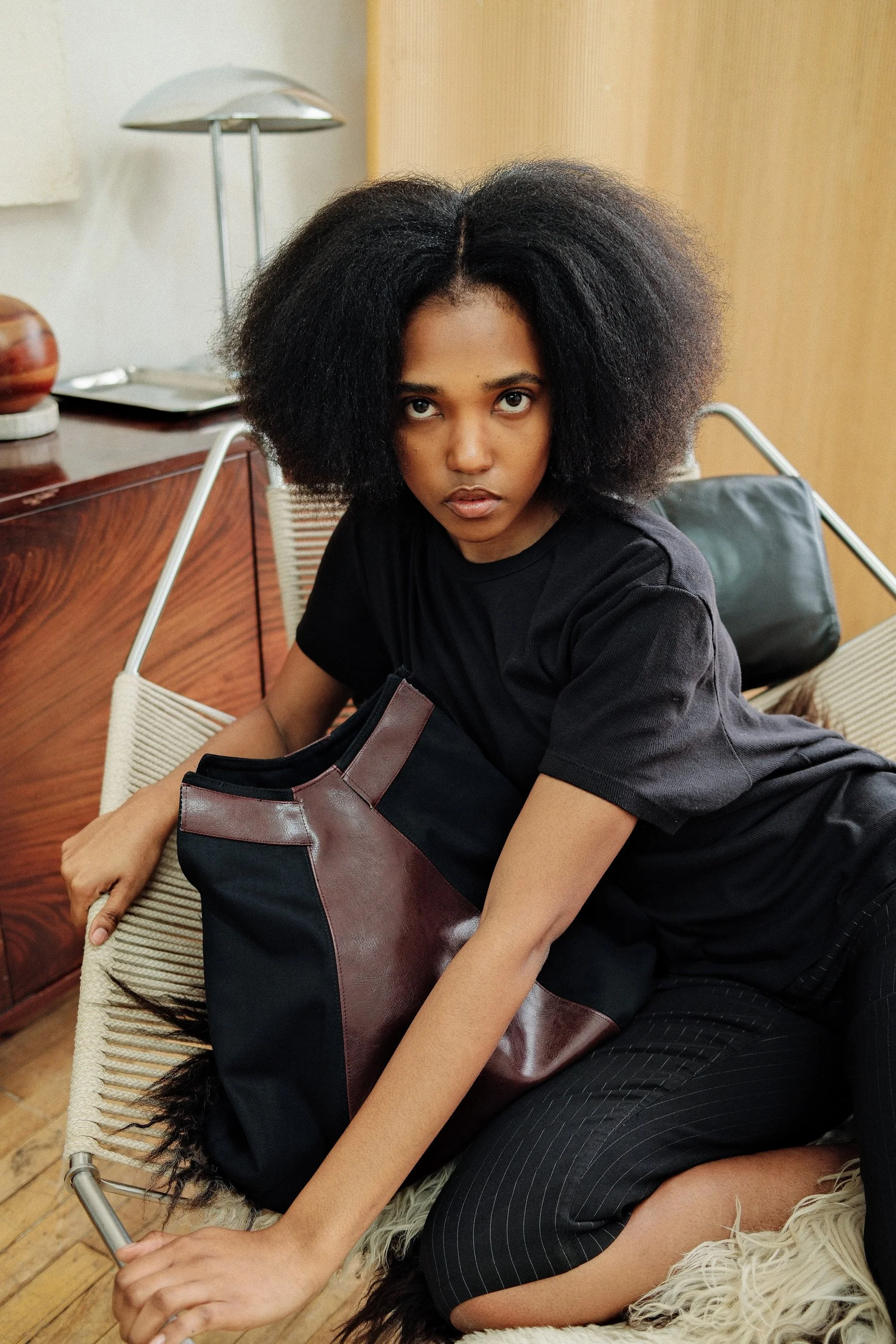 A young woman with natural, voluminous black hair sitting on a wicker chair in a room with wooden flooring and a wooden cabinet. She is wearing a black t-shirt and striped black pants, holding a phone in her right hand, and resting a large black and 
