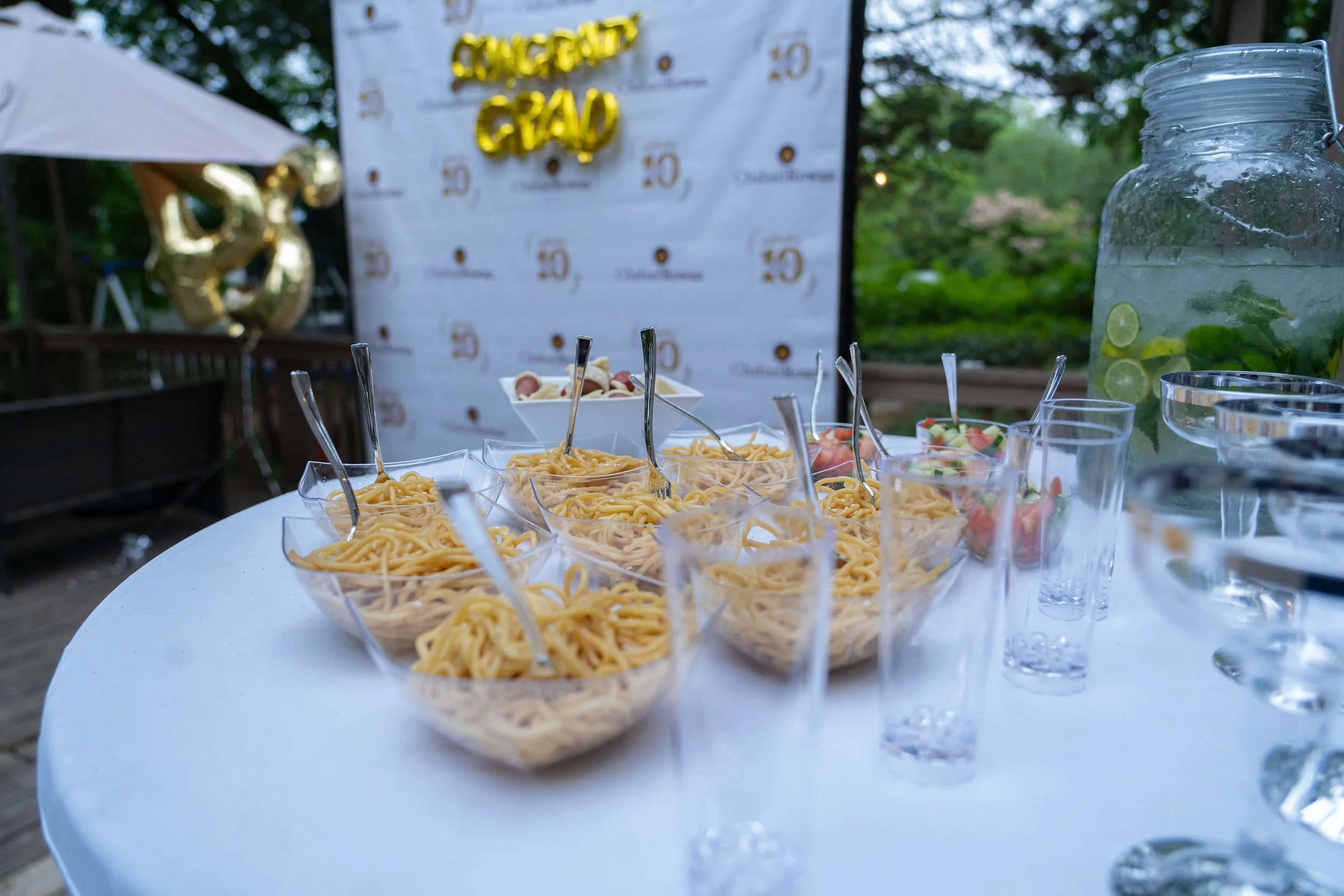A table with clear plastic bowls of spaghetti, with various toppings and sauces. In the background, there is a banner with gold text and some gold balloons, and a jar with lime slices and mint leaves, suggesting an outdoor celebration or event.