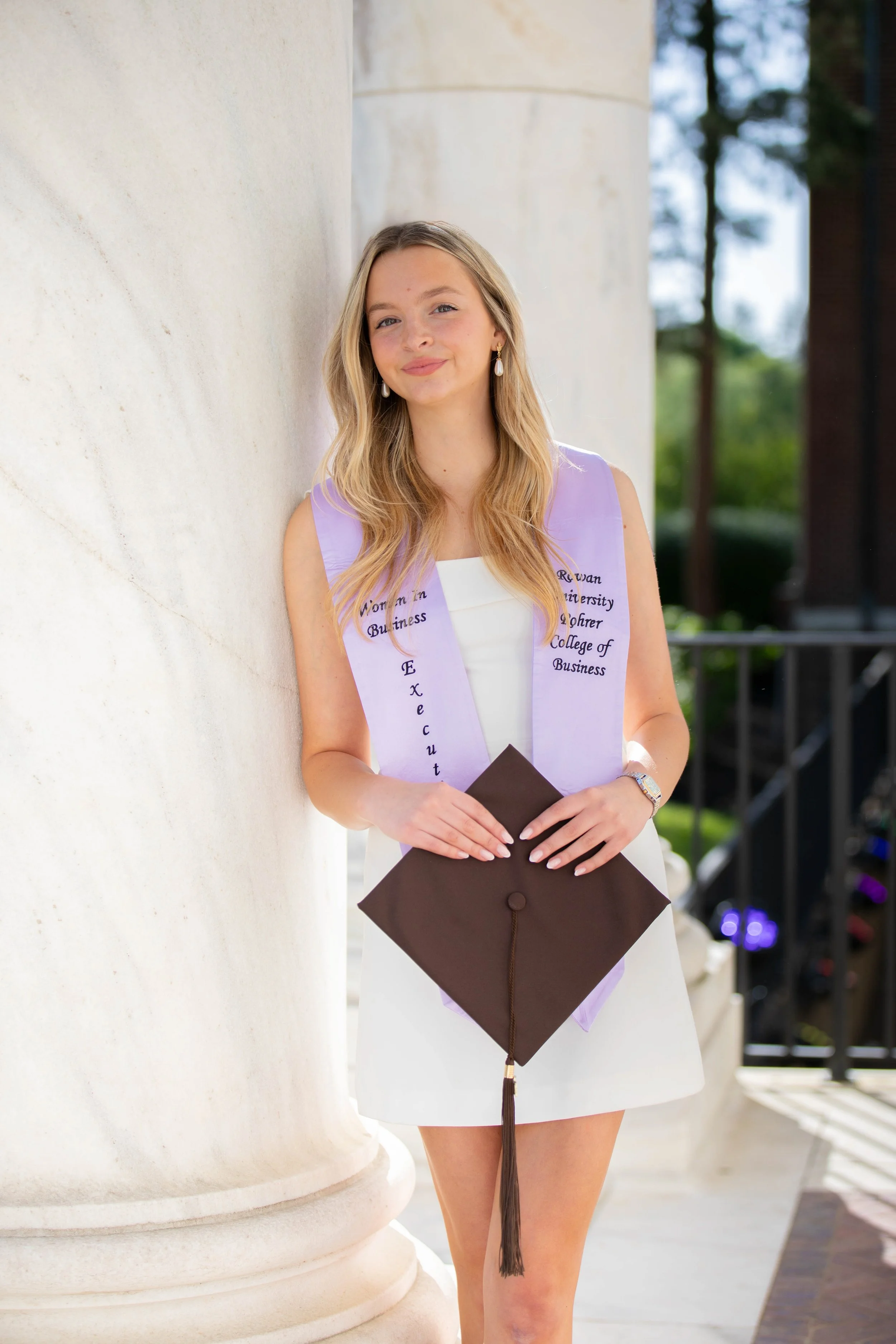 Young woman wearing a graduation stole and holding a diploma and cap, standing outdoors near a large white column.