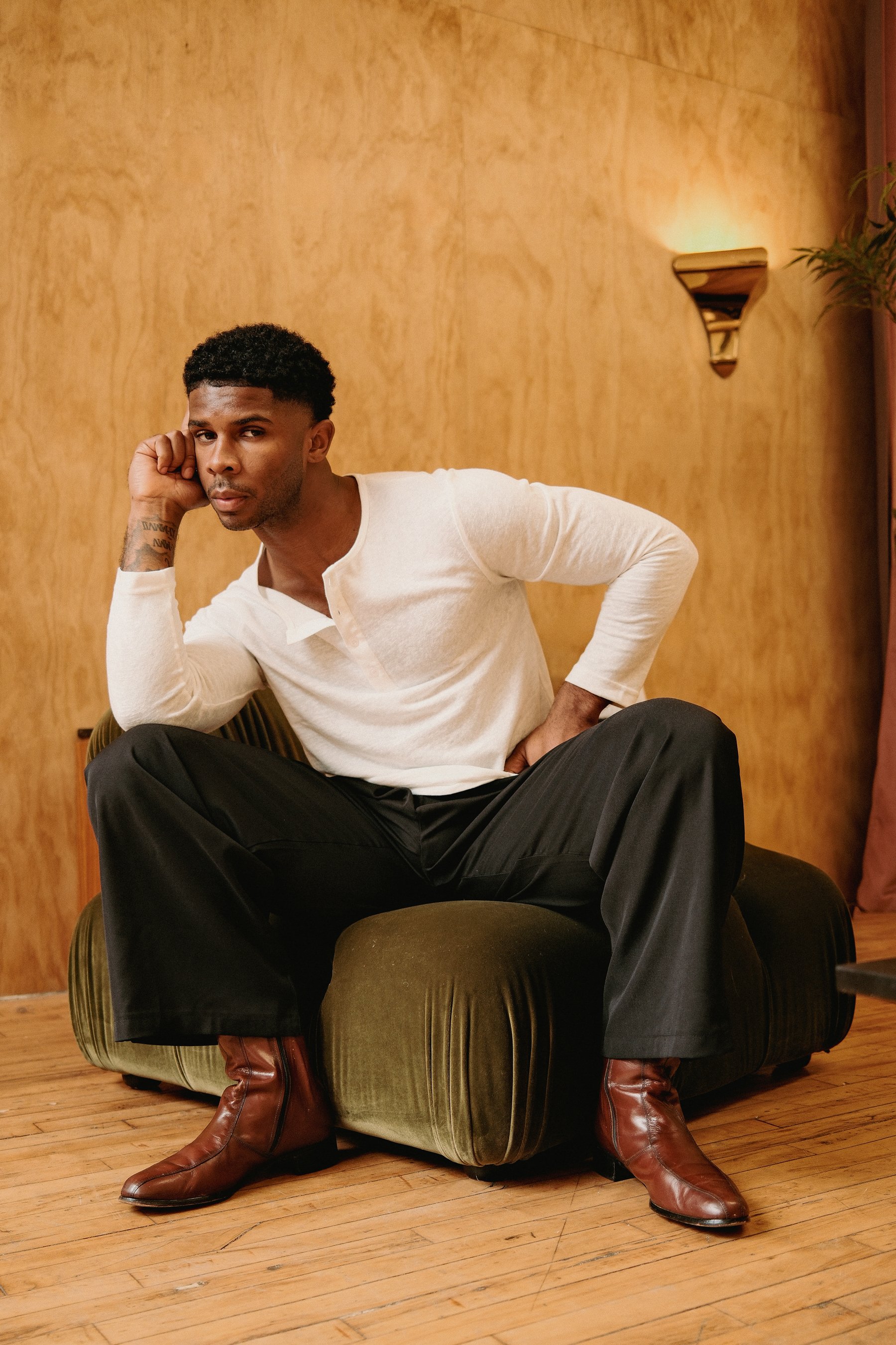 A man sitting on a vintage green velvet armchair, leaning forward slightly, with his right hand resting on his face and looking directly at the camera. He is wearing a white long-sleeve shirt, black pants, and brown leather boots. The background feat