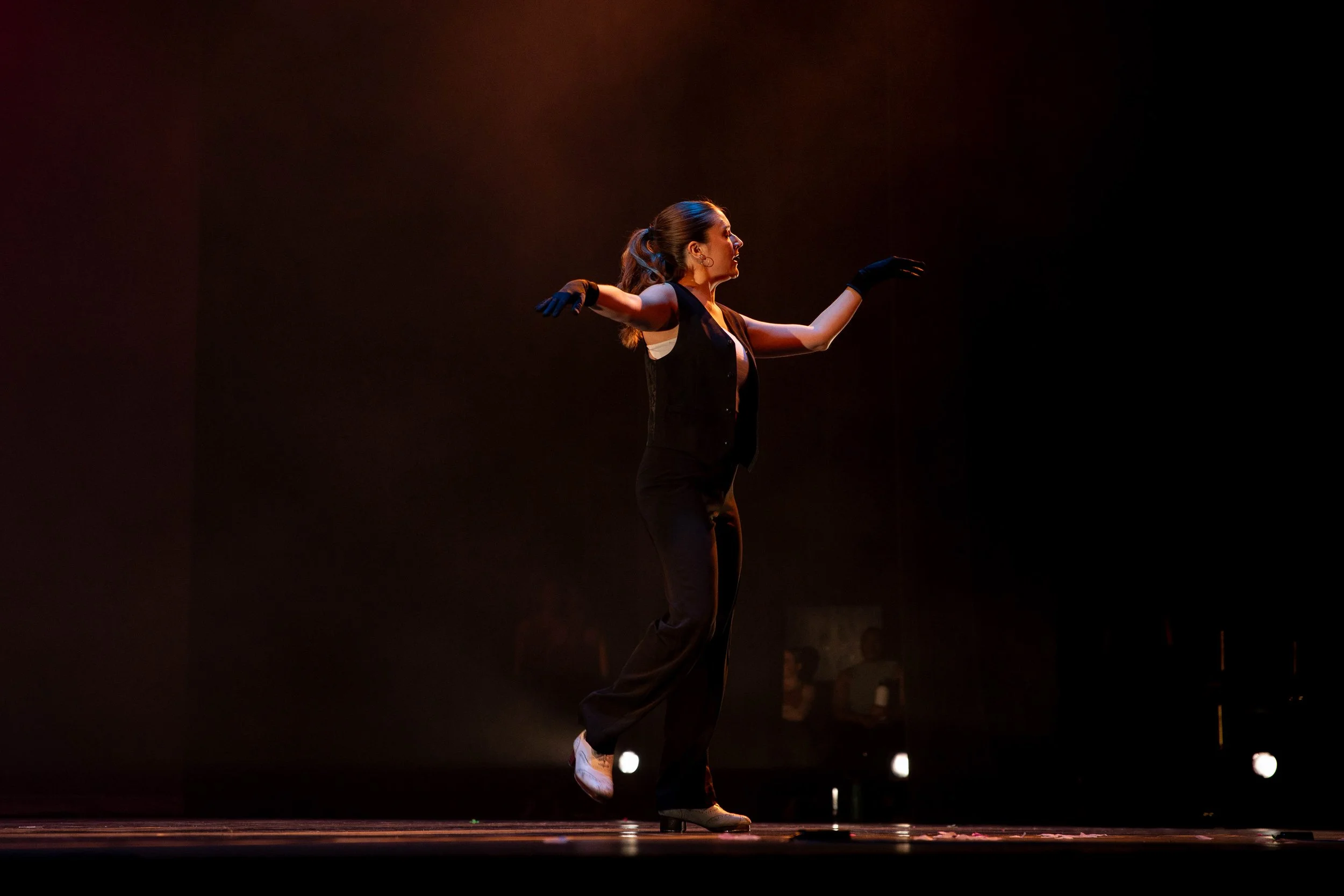 A female dancer performing on stage with her arms extended, wearing a black sleeveless top, black pants, black gloves, and white shoes, under warm stage lighting against a dark background.