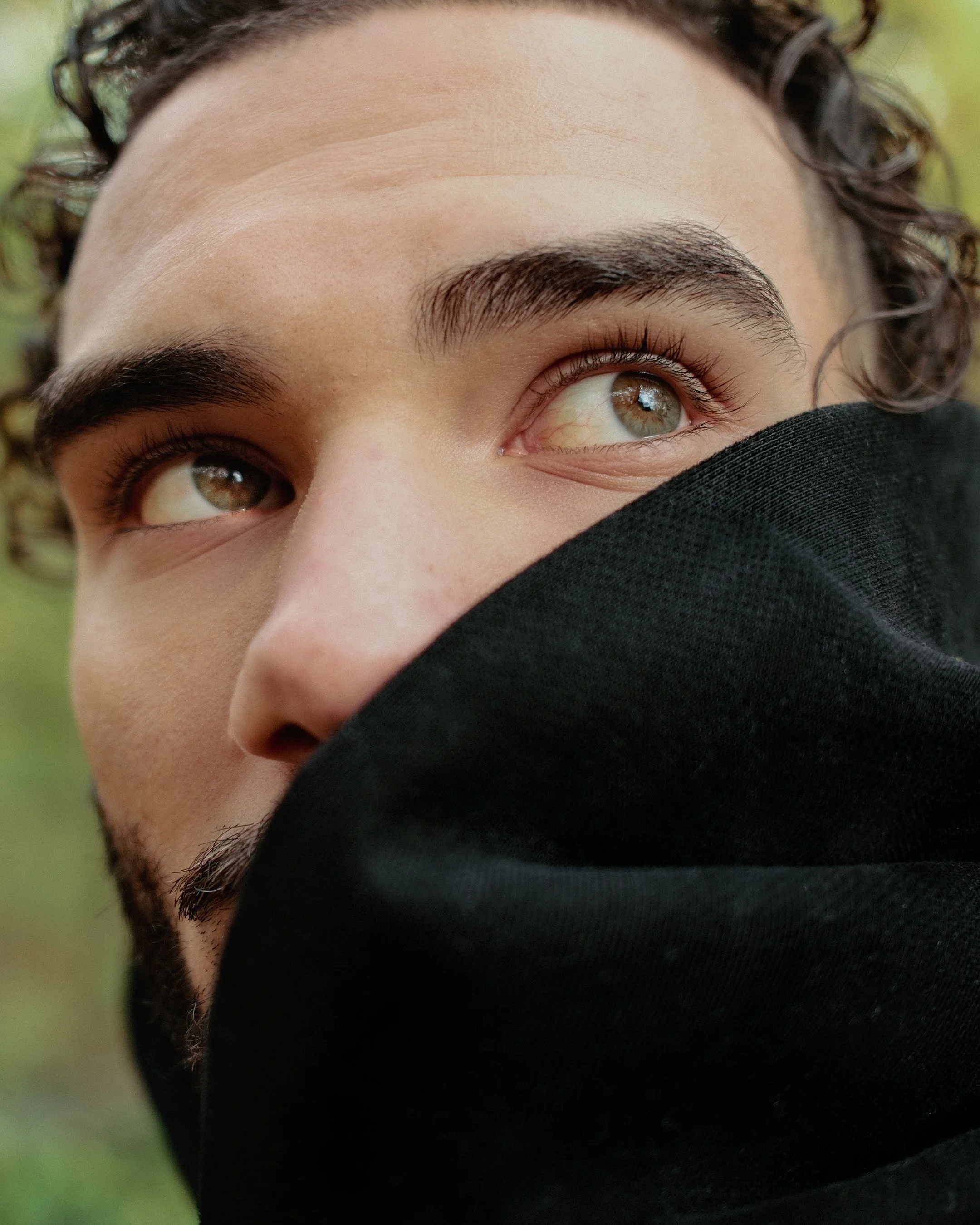 Close-up of a person's face with close-set brown eyes and thick eyebrows, partially covered by a black cloth or mask, with curly hair on the side and a blurred green background.