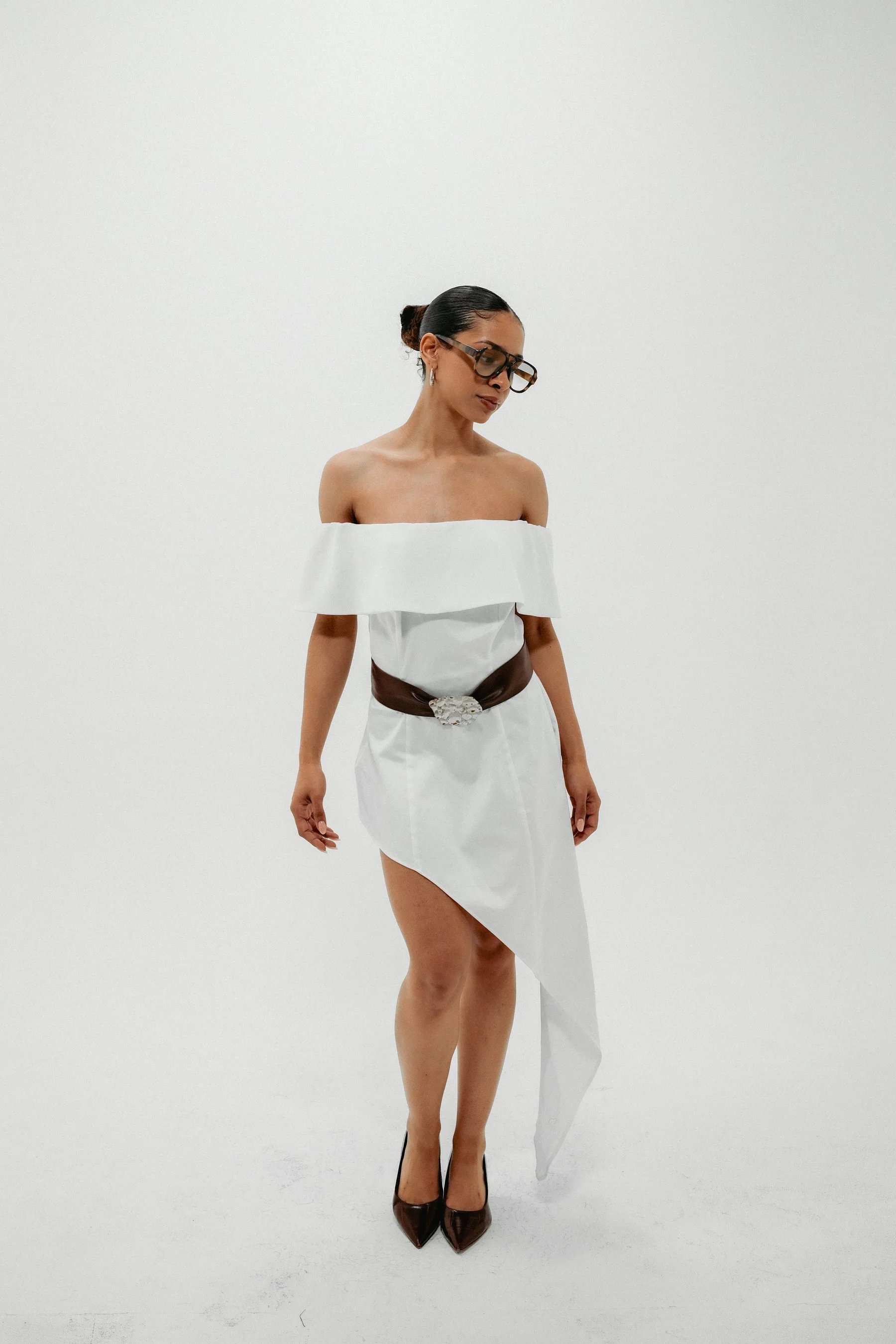 A woman with dark hair styled in a bun and wearing large sunglasses, a white off-the-shoulder dress with a slit, and a brown belt, stands against a plain white background.