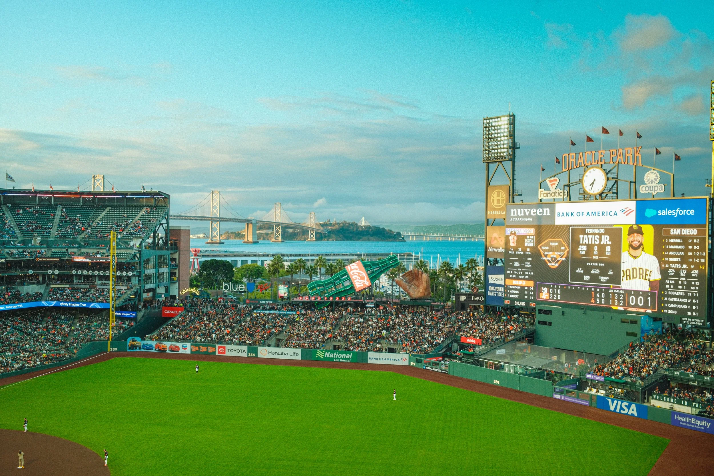 Scenes from Oracle Park baseball stadium in San Francisco during a game with a view of the bay and the San Francisco-Oakland Bay Bridge in the background. The stadium is filled with spectators, and the scoreboard displays player statistics.