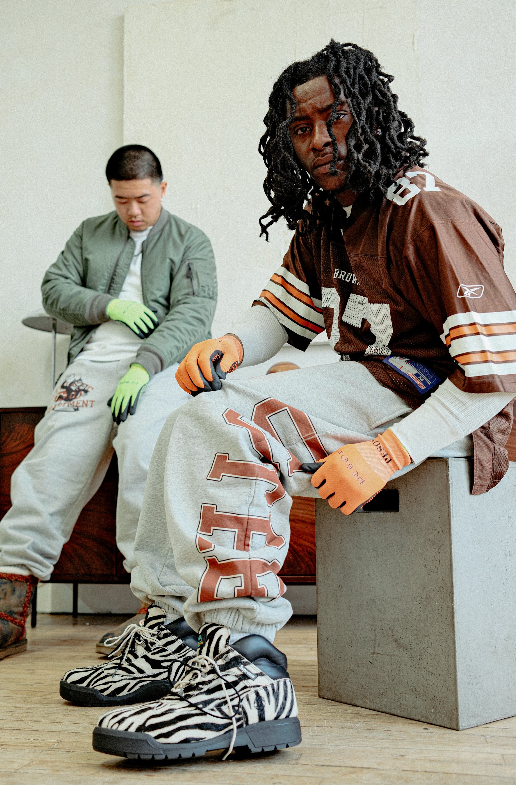Two young men sitting on a bench in a room with a plain white wall. One man is looking down, wearing a green bomber jacket and white pants, with neon green gloves. The other man in the foreground is wearing a brown sports jersey and grey sweatpants, 