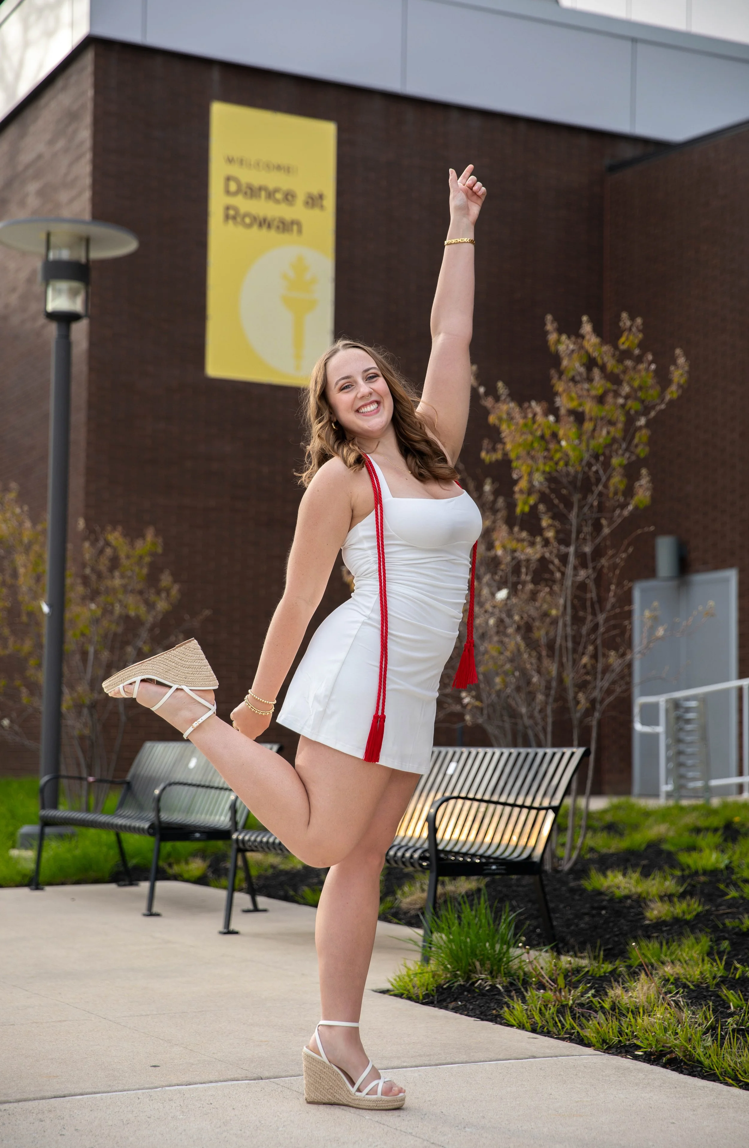 A young woman in a white dress and wedge sandals is jumping with her knee bent and one arm raised, smiling brightly outdoors near a bench and small trees, with a brick building and a yellow sign in the background that reads "Welcome! Dance at Rowan".